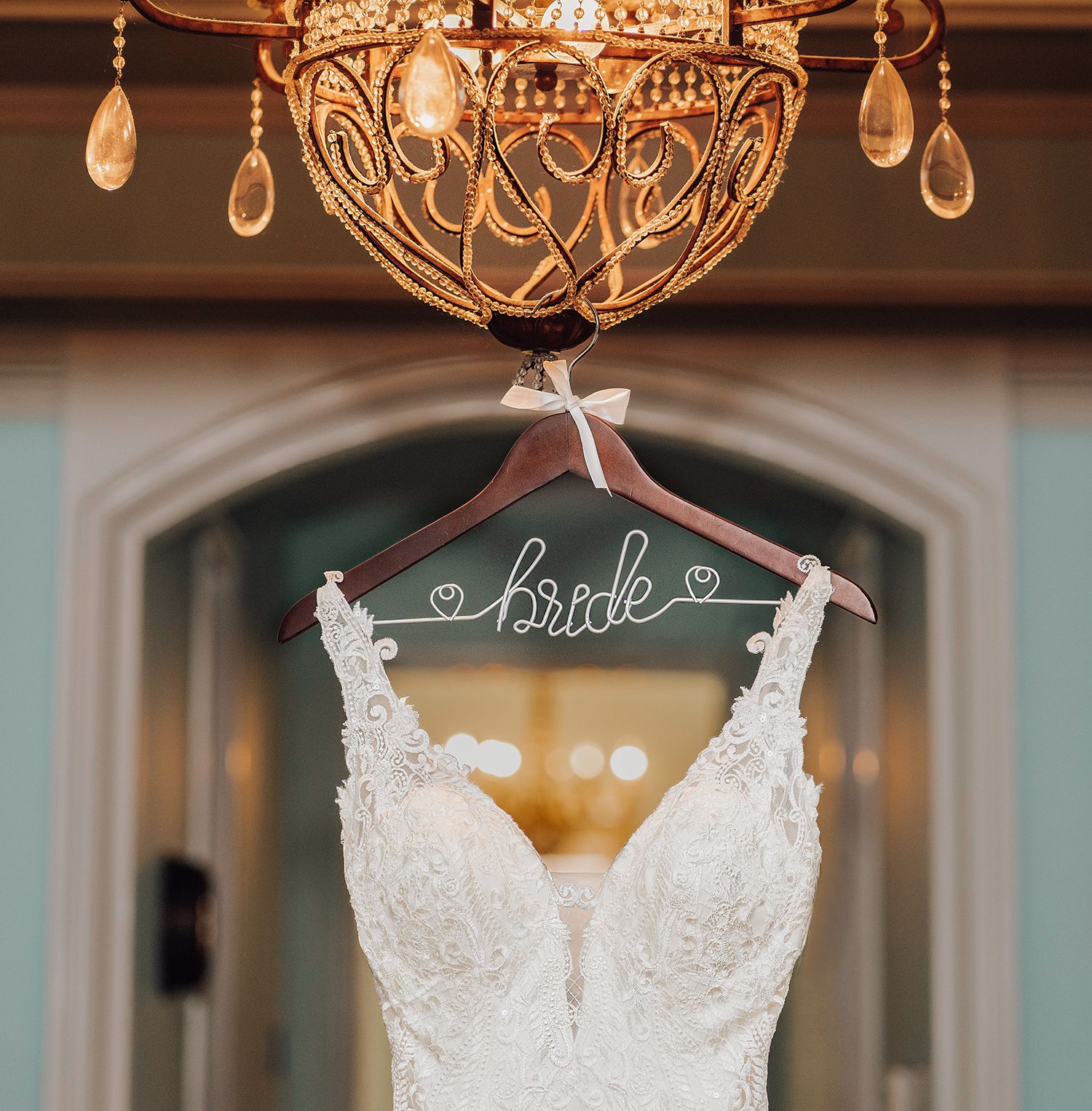 A white lace wedding dress with beaded details hangs from a crystal chandelier in an elegant room.