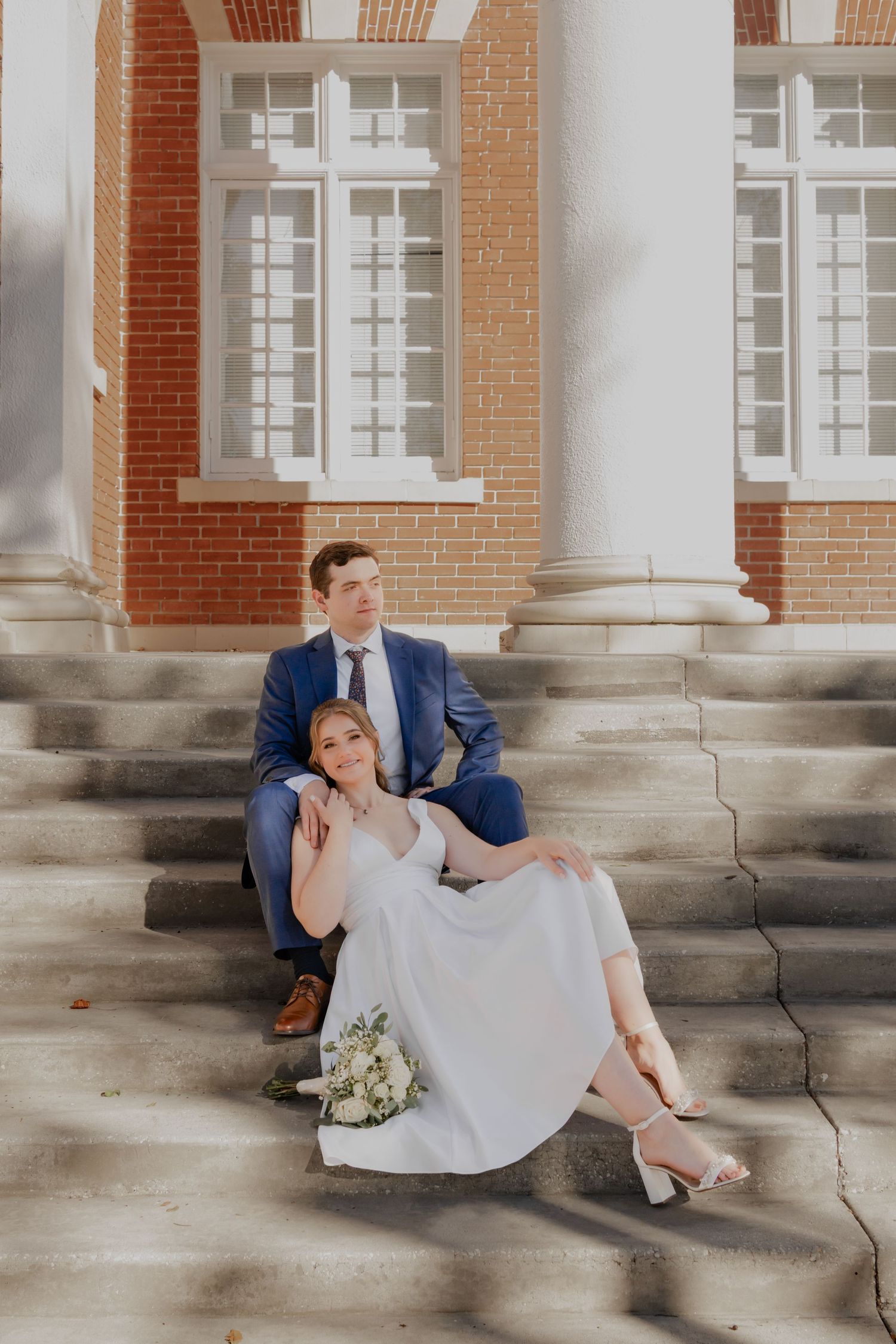 Bride and groom sitting on steps of brick building, woman in white dress with bouquet, man in blue suit.