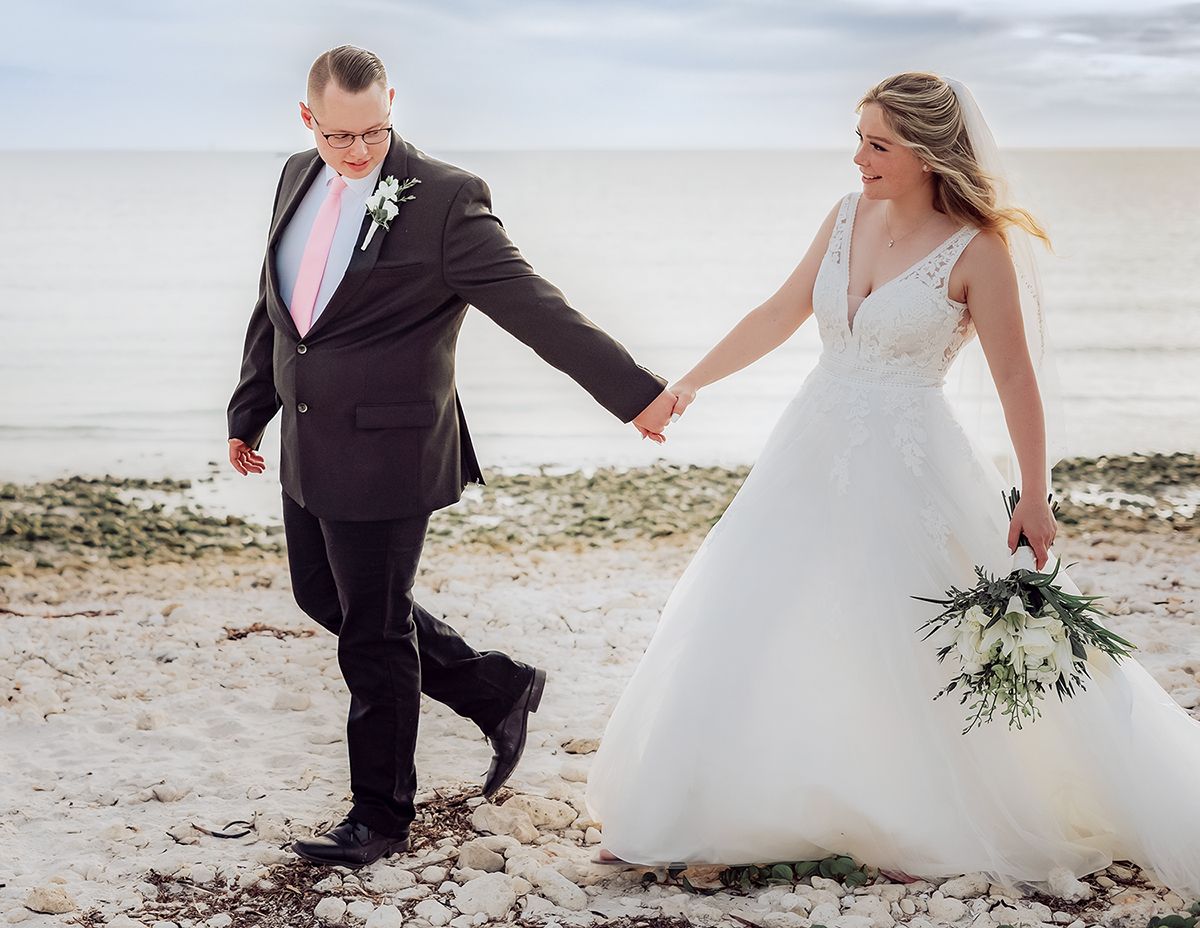 A couple in wedding attire walks hand in hand along a beach, with the bride holding a delicate white bouquet.
