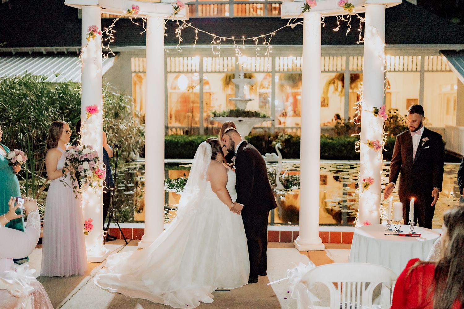 A romantic evening wedding ceremony takes place under an illuminated white gazebo with decorative floral arrangements.