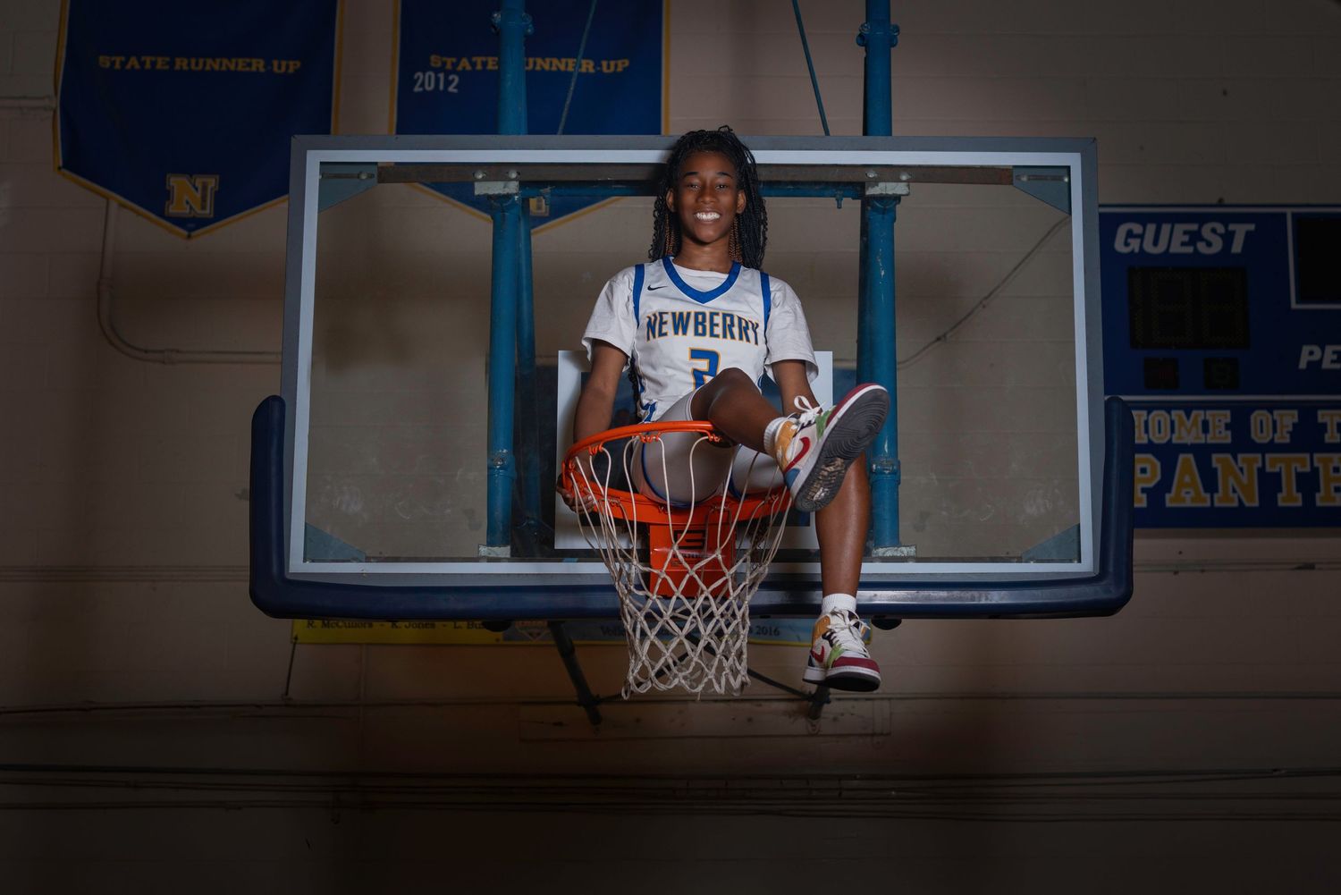Basketball player sitting in hoop wearing Newberry jersey, smiling in gymnasium.