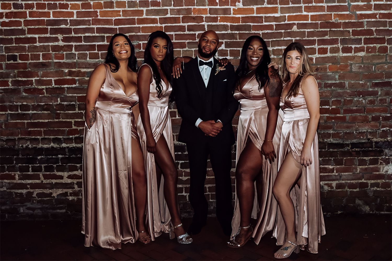 A group in formal attire poses together against a rustic brick wall, wearing satin gowns and a black tuxedo for wedding photographer in Jax.