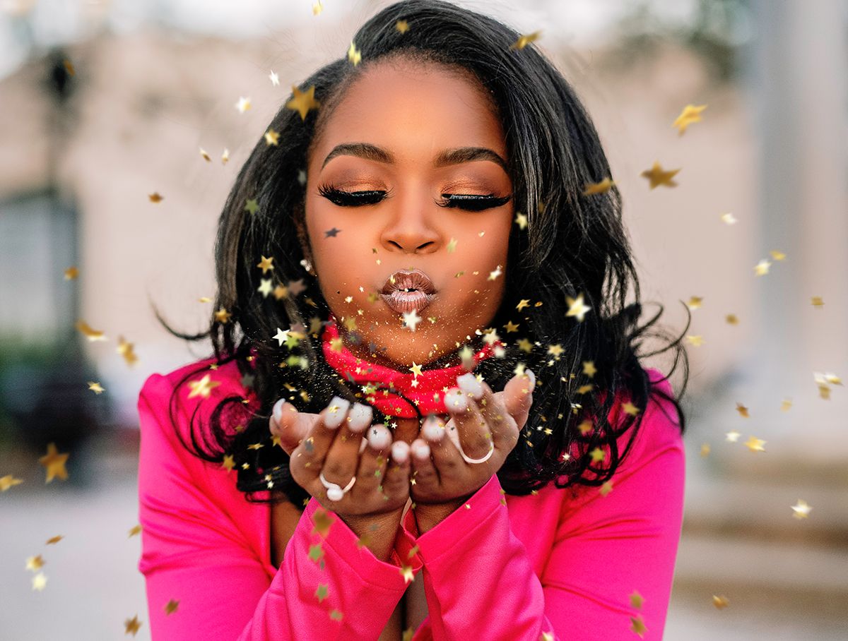 Woman in pink blowing gold star confetti from her hands, celebrating with joy outdoors.