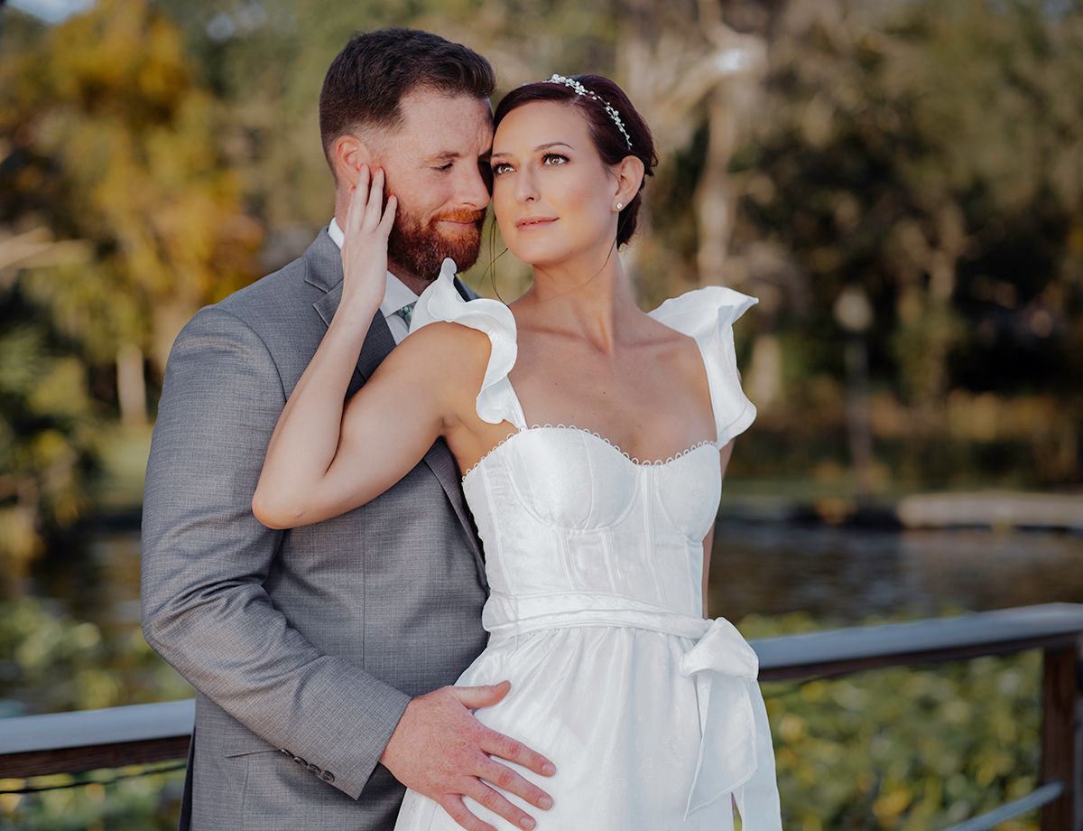 Bride in white ruffled gown and groom in gray suit share romantic embrace outdoors near lush greenery.