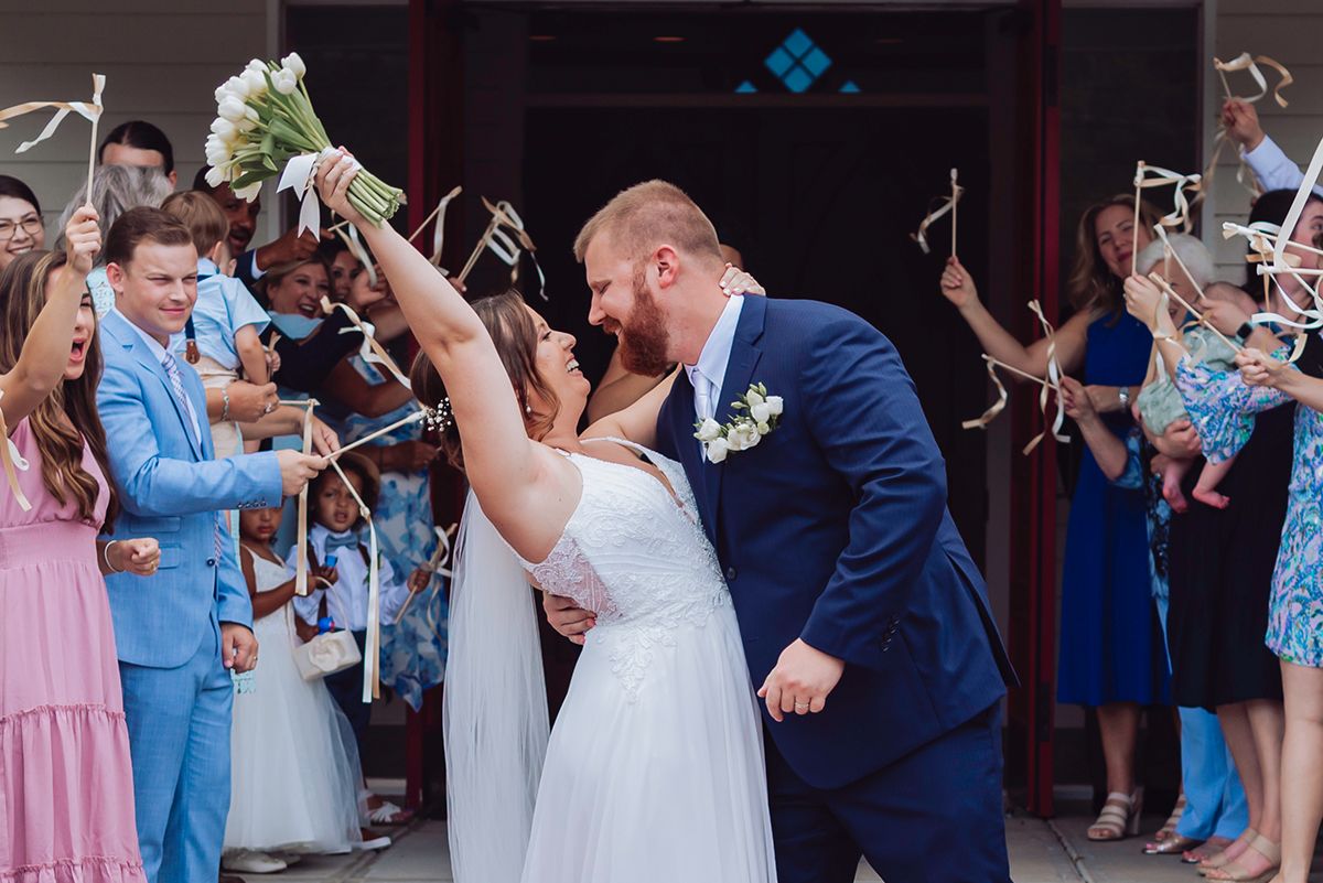 Newlywed couple sharing a kiss while guests wave ribbon wands during a joyful outdoor wedding ceremony exit.
