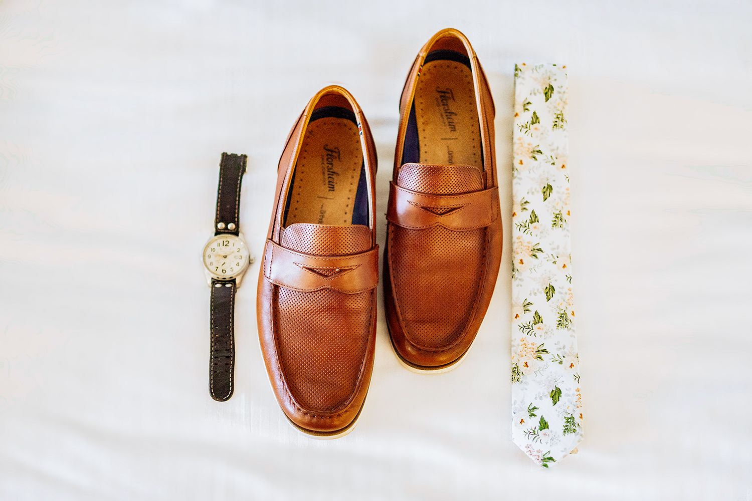 Brown leather penny loafers and a watch arranged on a white surface with delicate floral accents groom details for wedding photography