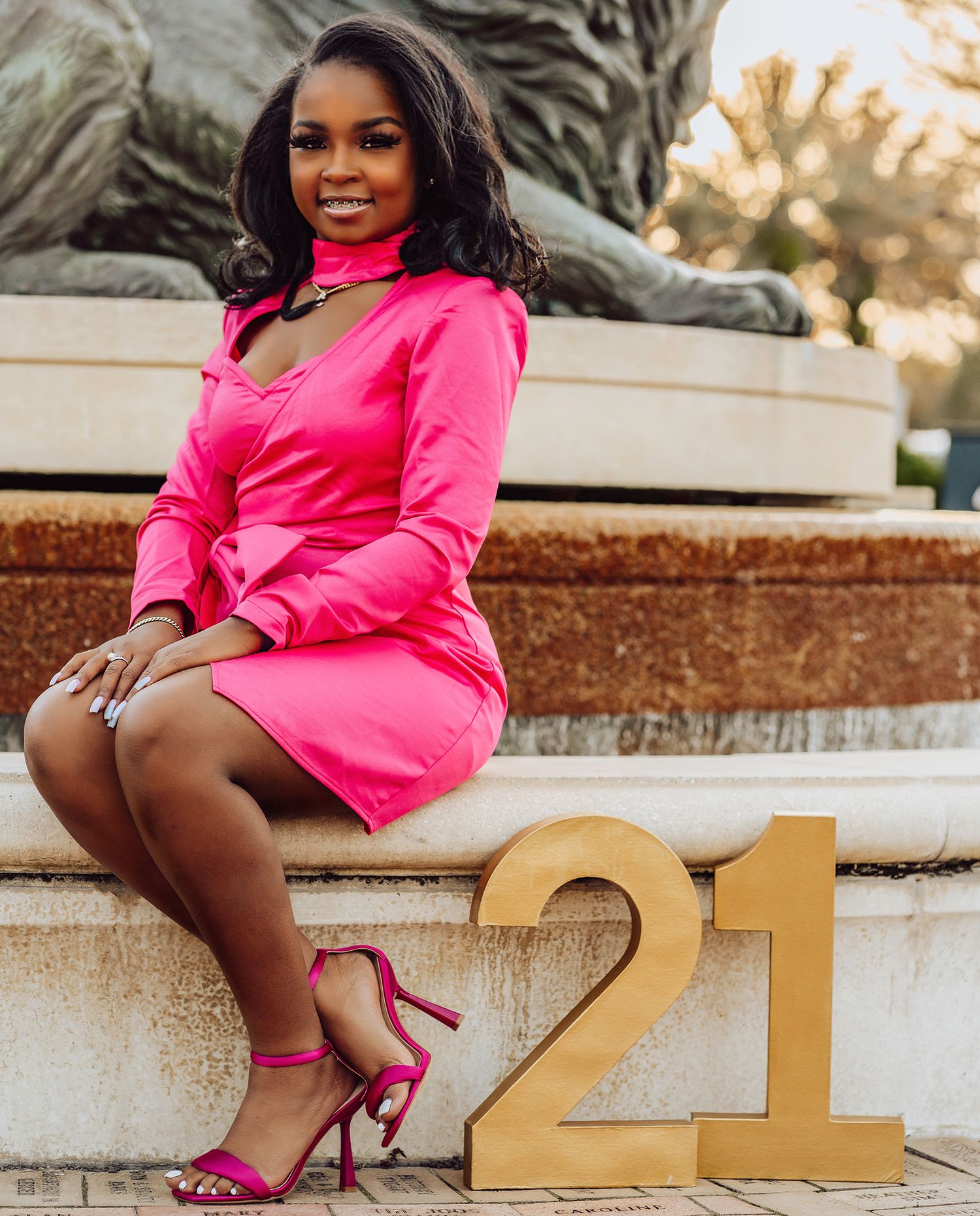 High school senior wearing bright pink outfit poses with large gold numbers for a high school graduation photoshoot at sunset.
