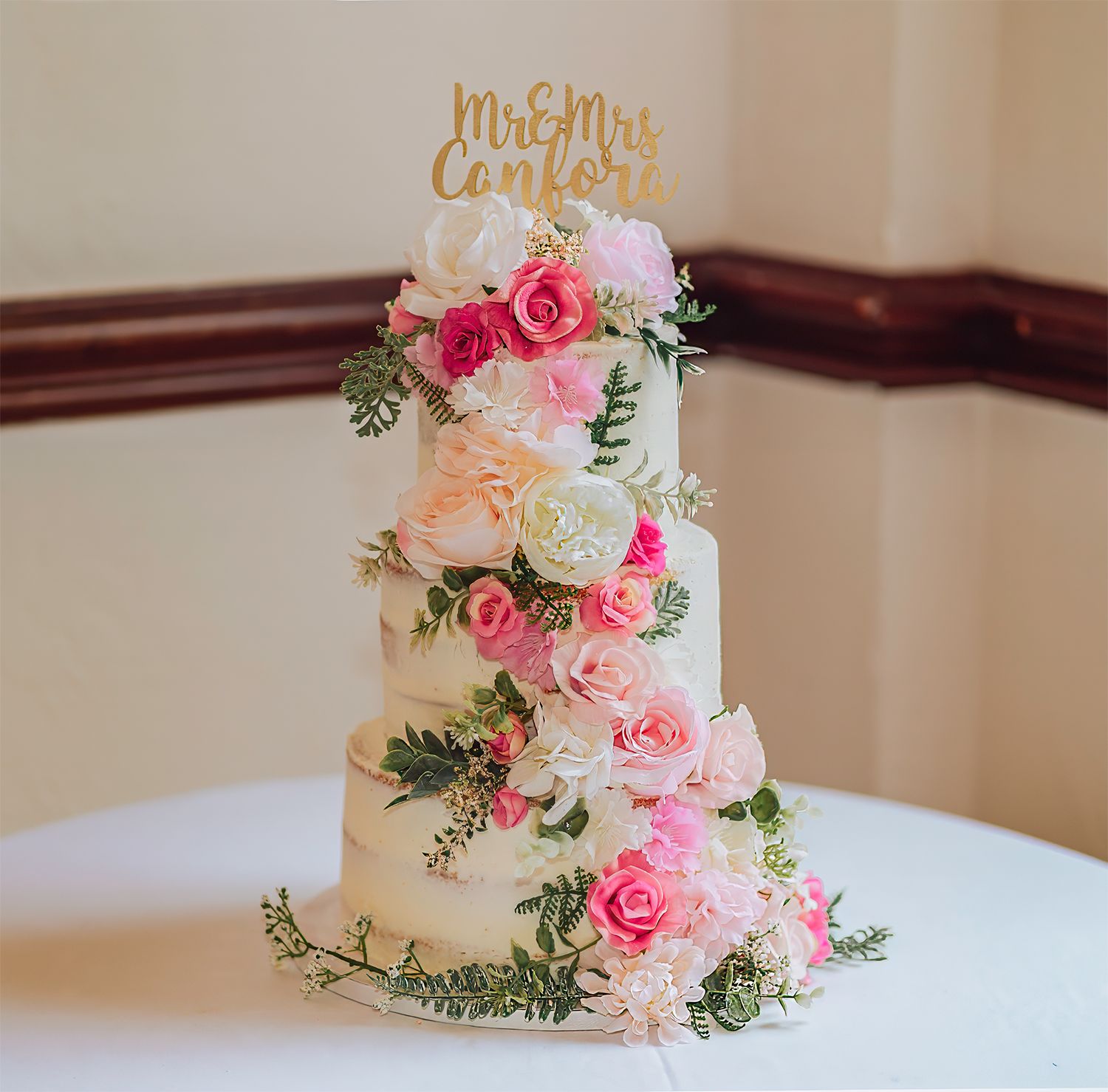 Elegant three-tier white wedding cake decorated with pink roses, greenery and a gold Mr and Mrs topper on white tablecloth.