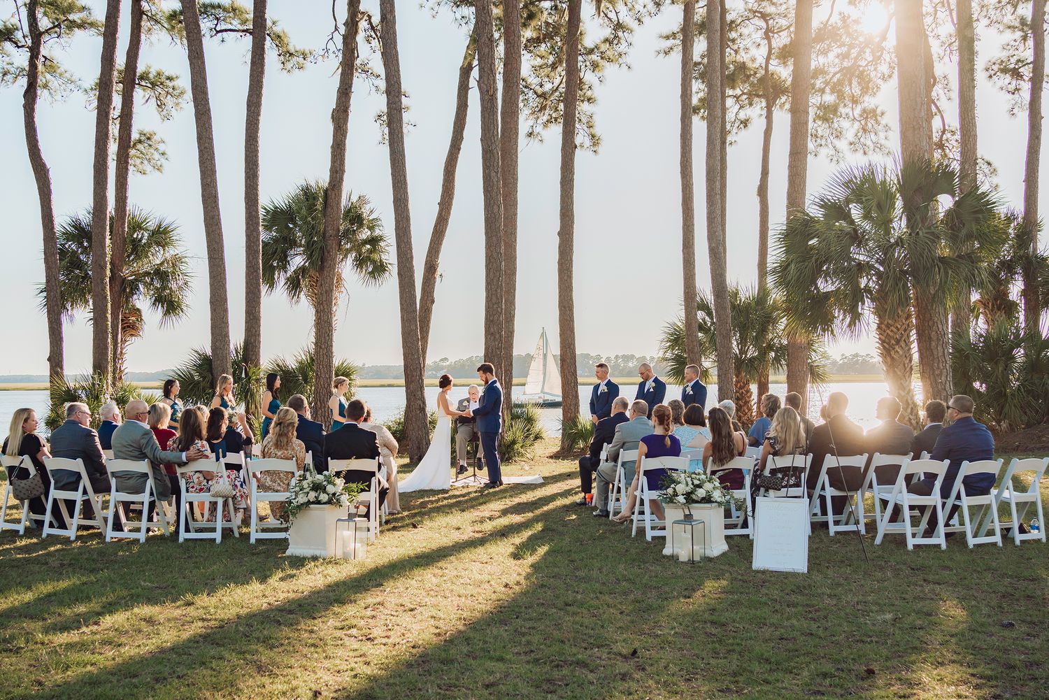 Outdoor wedding ceremony setup with white chairs among palm trees at sunset.