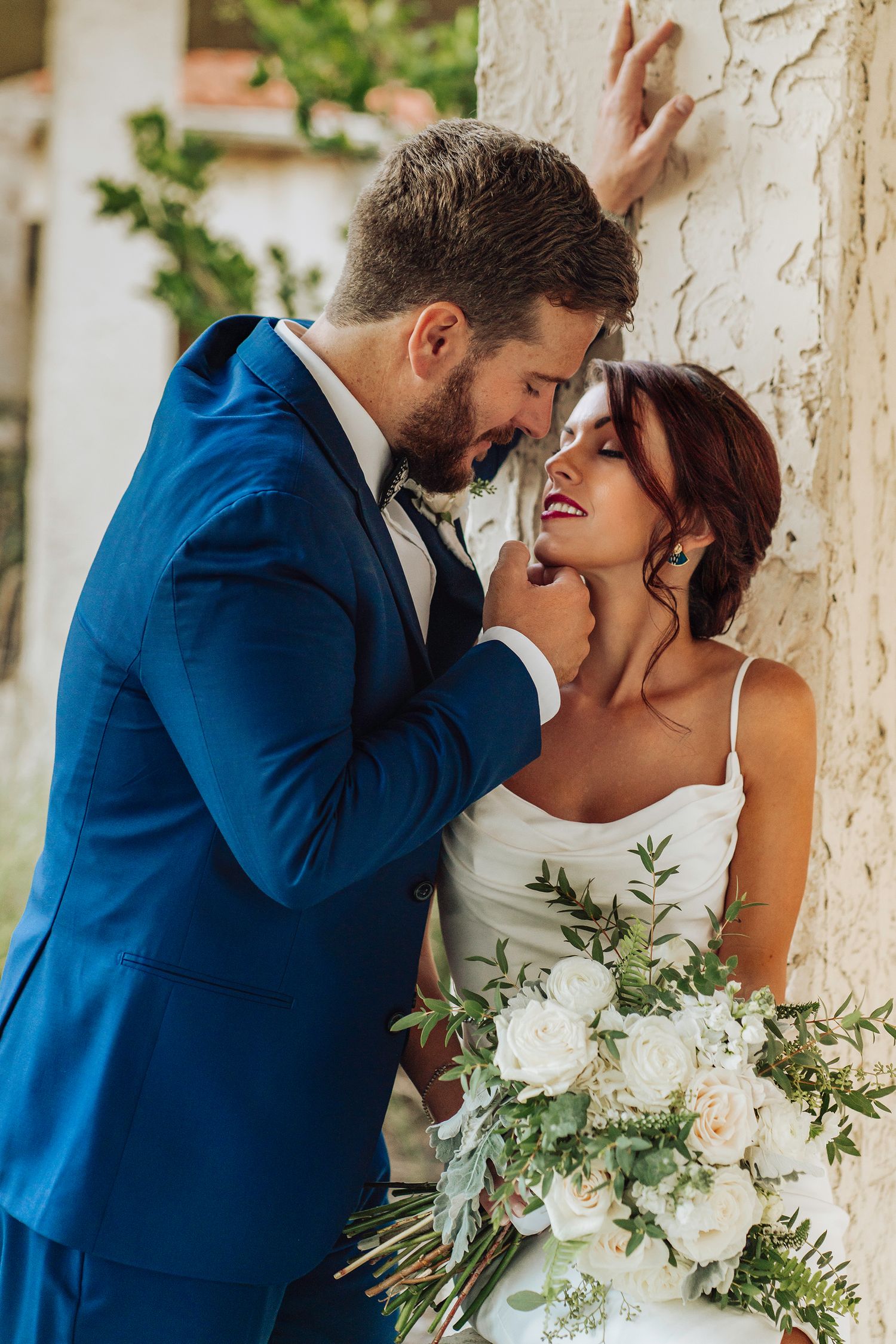 Romantic wedding couple in blue suit and white dress share intimate moments against rustic wall with floral bouquet.