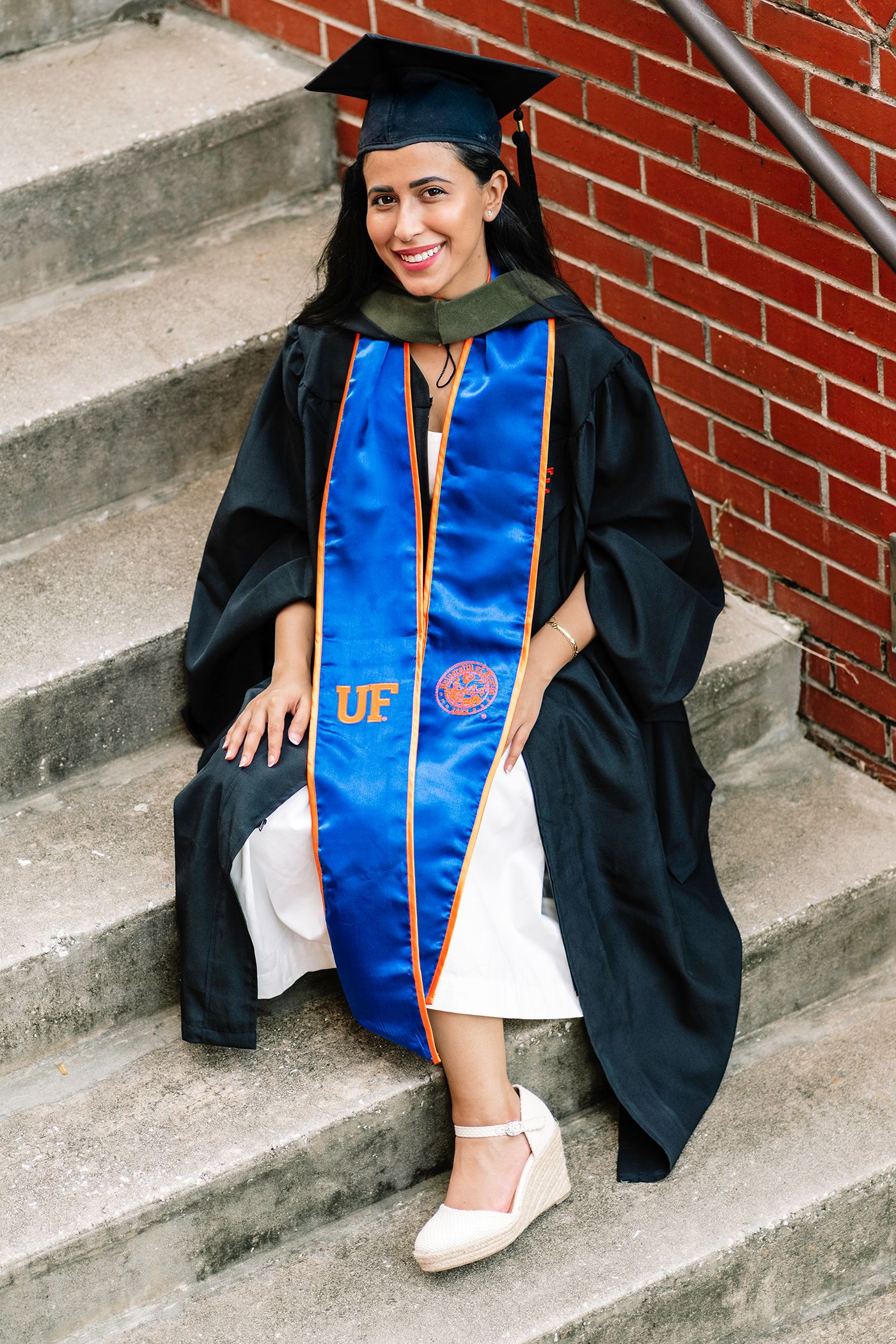Graduate in cap and gown with UF blue and orange stole sits on concrete steps by brick wall wearing white dress and flats.