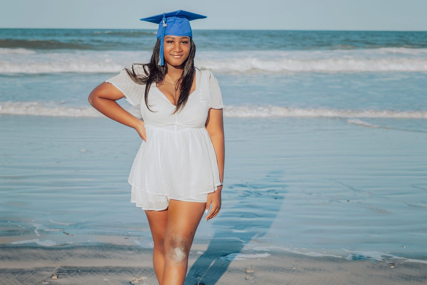 Young woman in white dress and blue graduation cap posing on a sunny beach with ocean waves behind her.