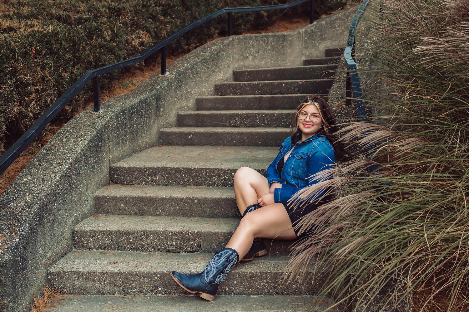 High school senior in blue denim jacket sits on concrete stairs at Oglebay Zoo in Wheeling, WV for senior photos.