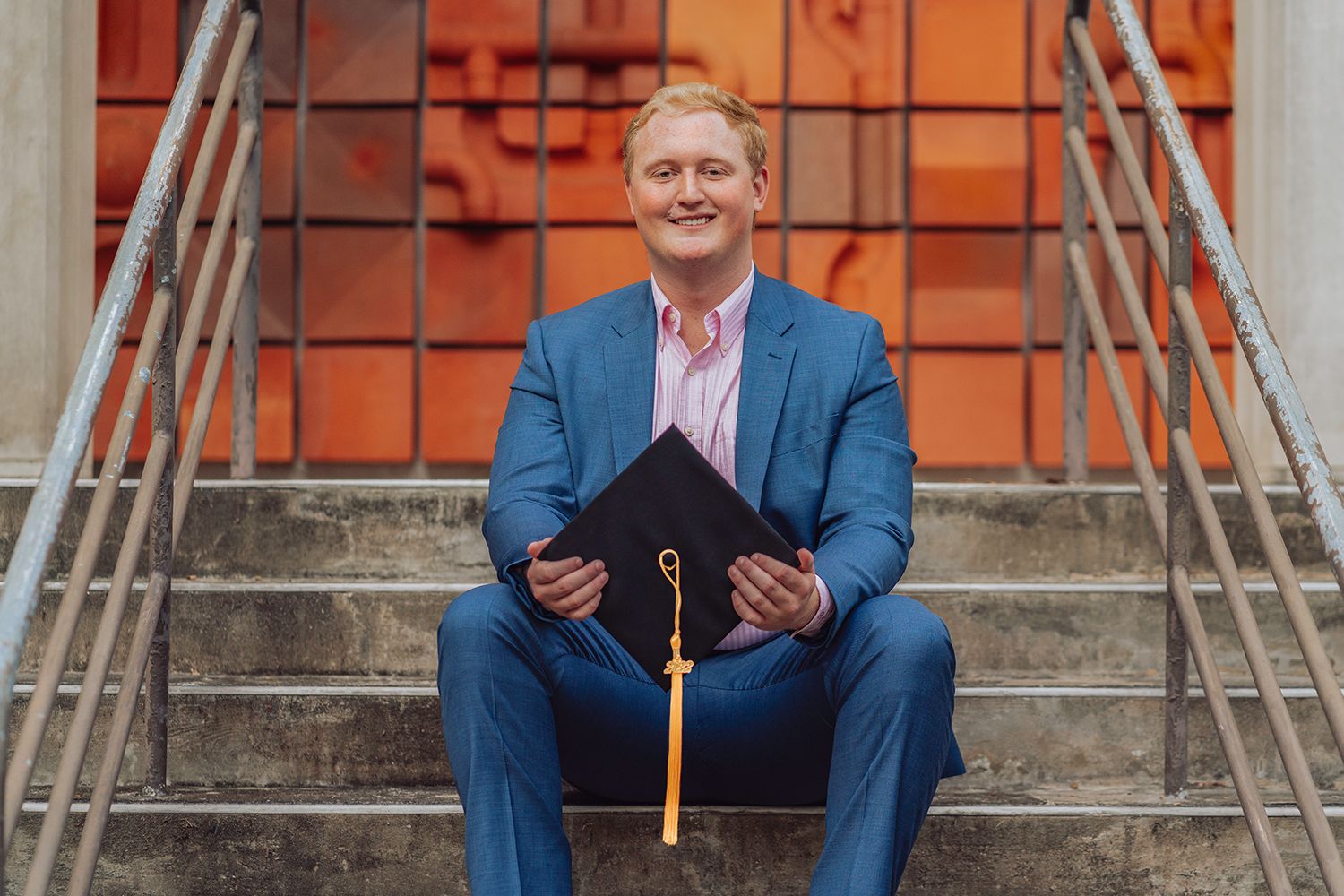 Graduate in blue suit sits on stone steps holding diploma and tassel while smiling at camera at University of Florida, Gainesville, Florida.