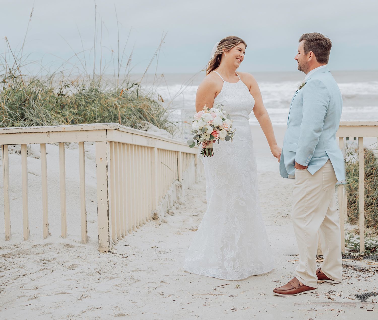 A couple in wedding attire shares a romantic moment on a wooden boardwalk leading to a sandy beach.