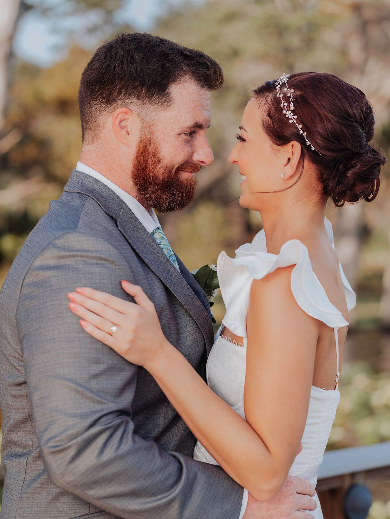 A bride and groom in wedding attire share a romantic moment, gazing into each other's eyes outdoors.