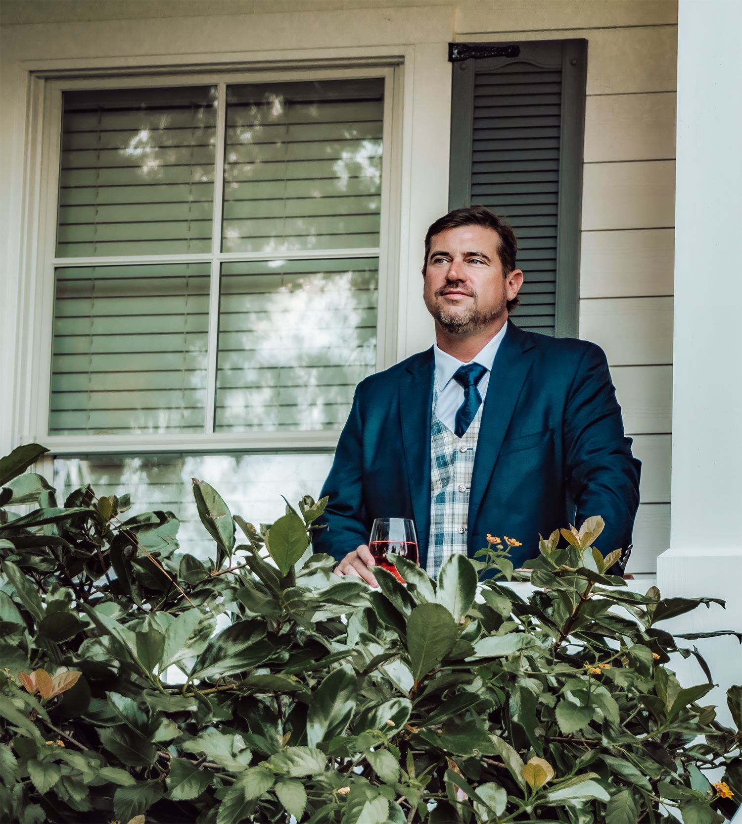 Professional in a navy suit stands on a front porch with white columns and green foliage in the background.