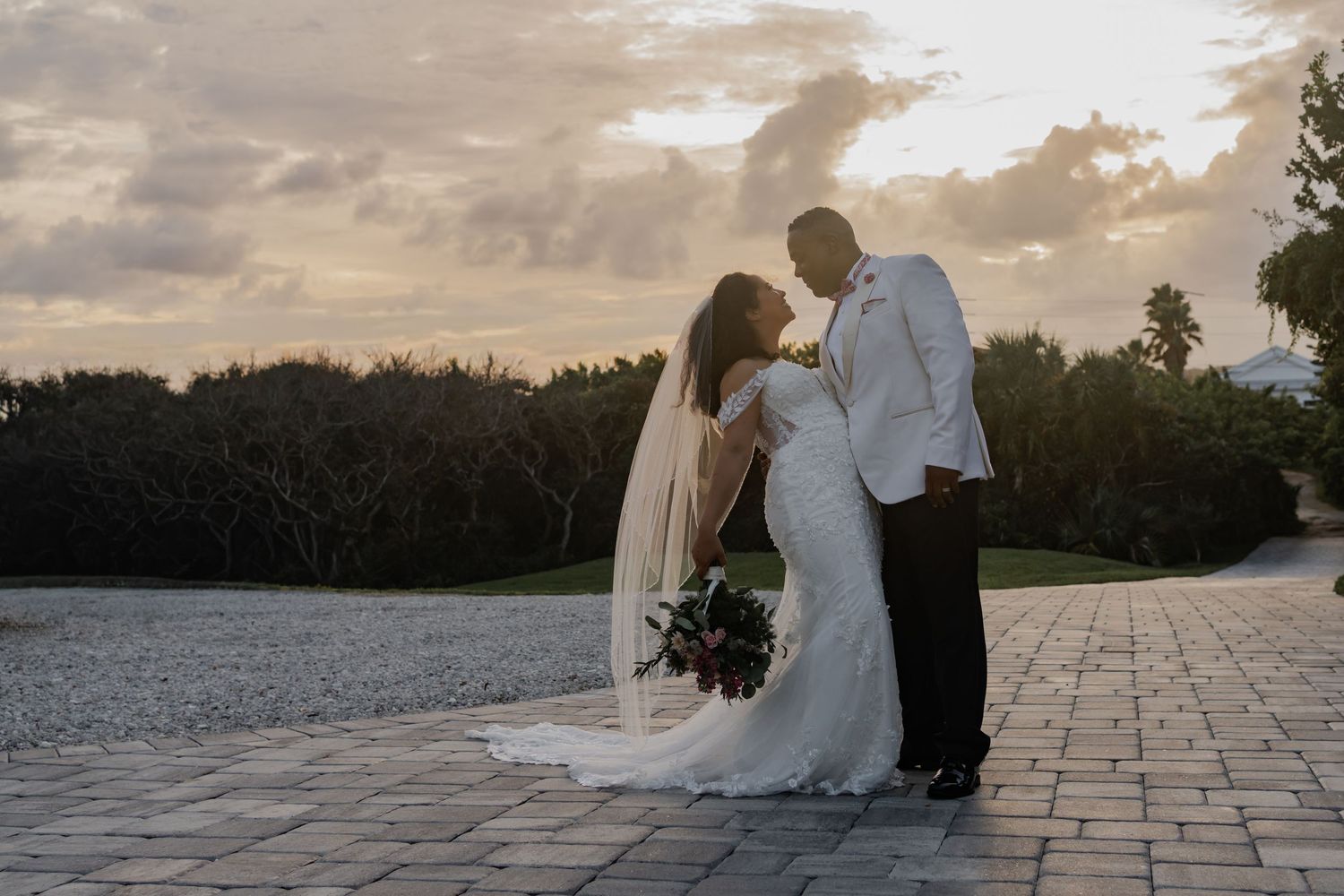 Bride and groom sharing a romantic sunset kiss on a stone patio, bride in white gown with veil holding bouquet.