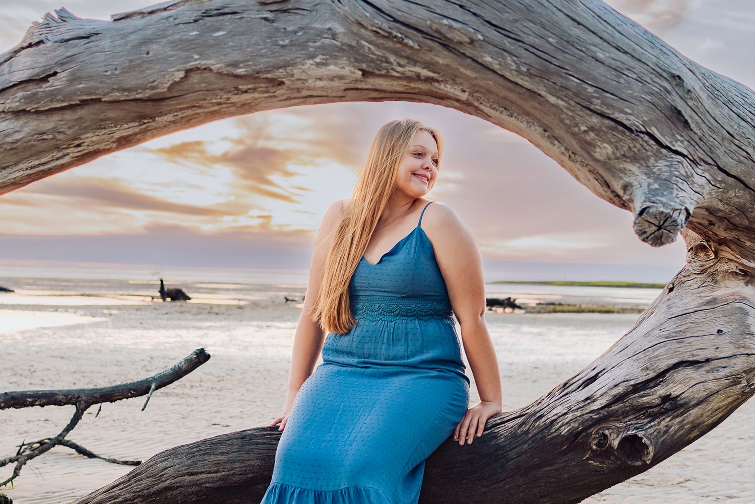High school senior  in blue dress poses on driftwood at beach during sunset for graduation photos in Chiefland, Florida