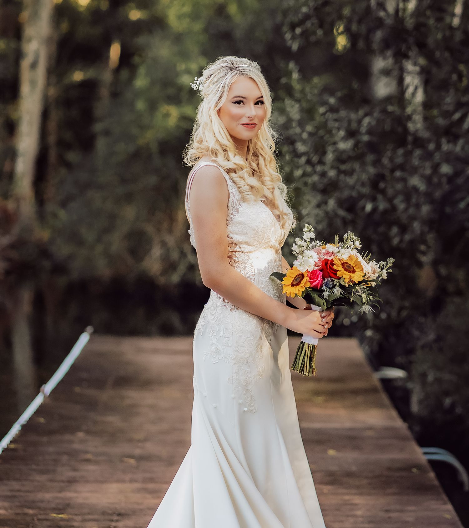 A bride in a white lace dress holds a vibrant sunflower bouquet while posing outdoors in a wooded setting.