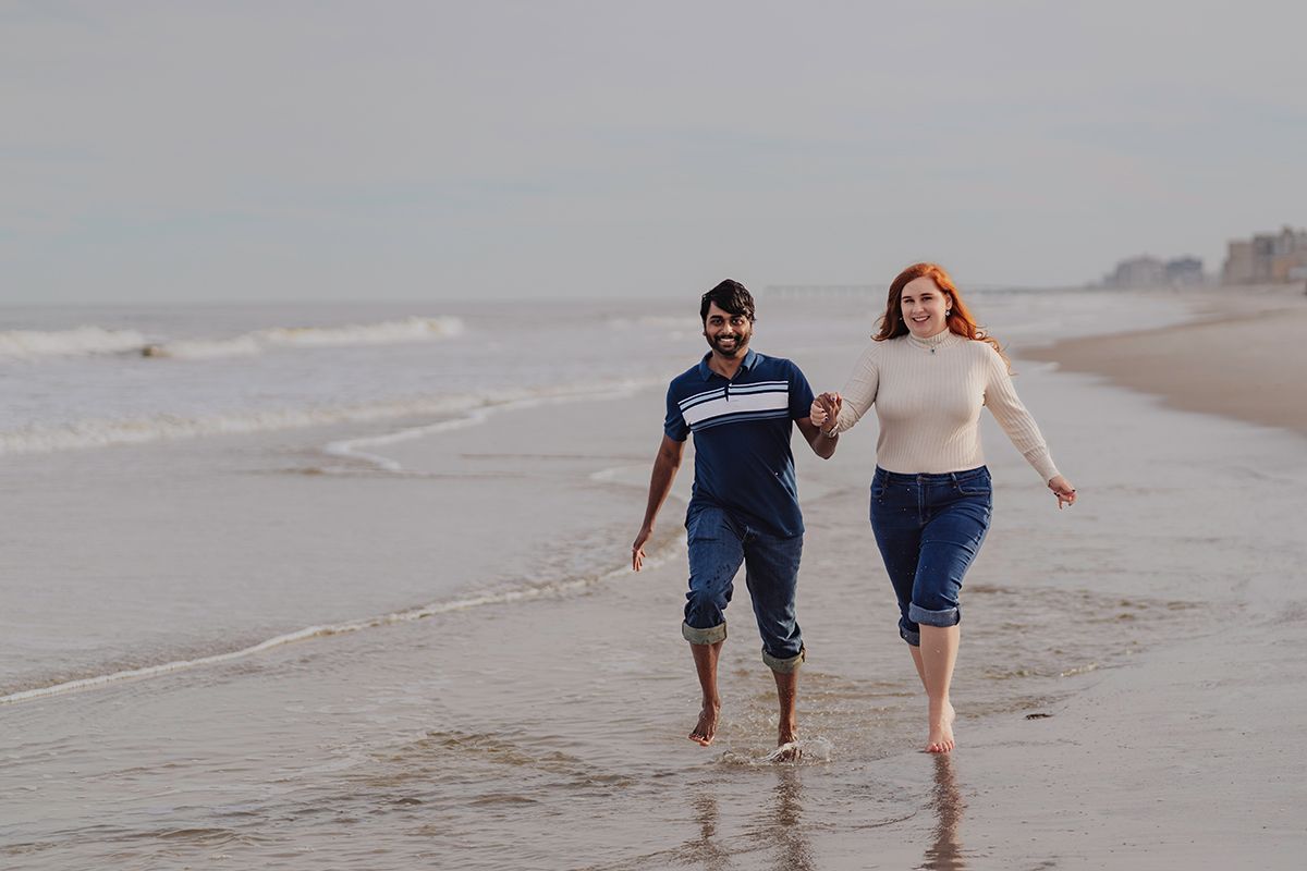 A couple holds hands and walks barefoot along a sandy beach shore with gentle ocean waves in the background.