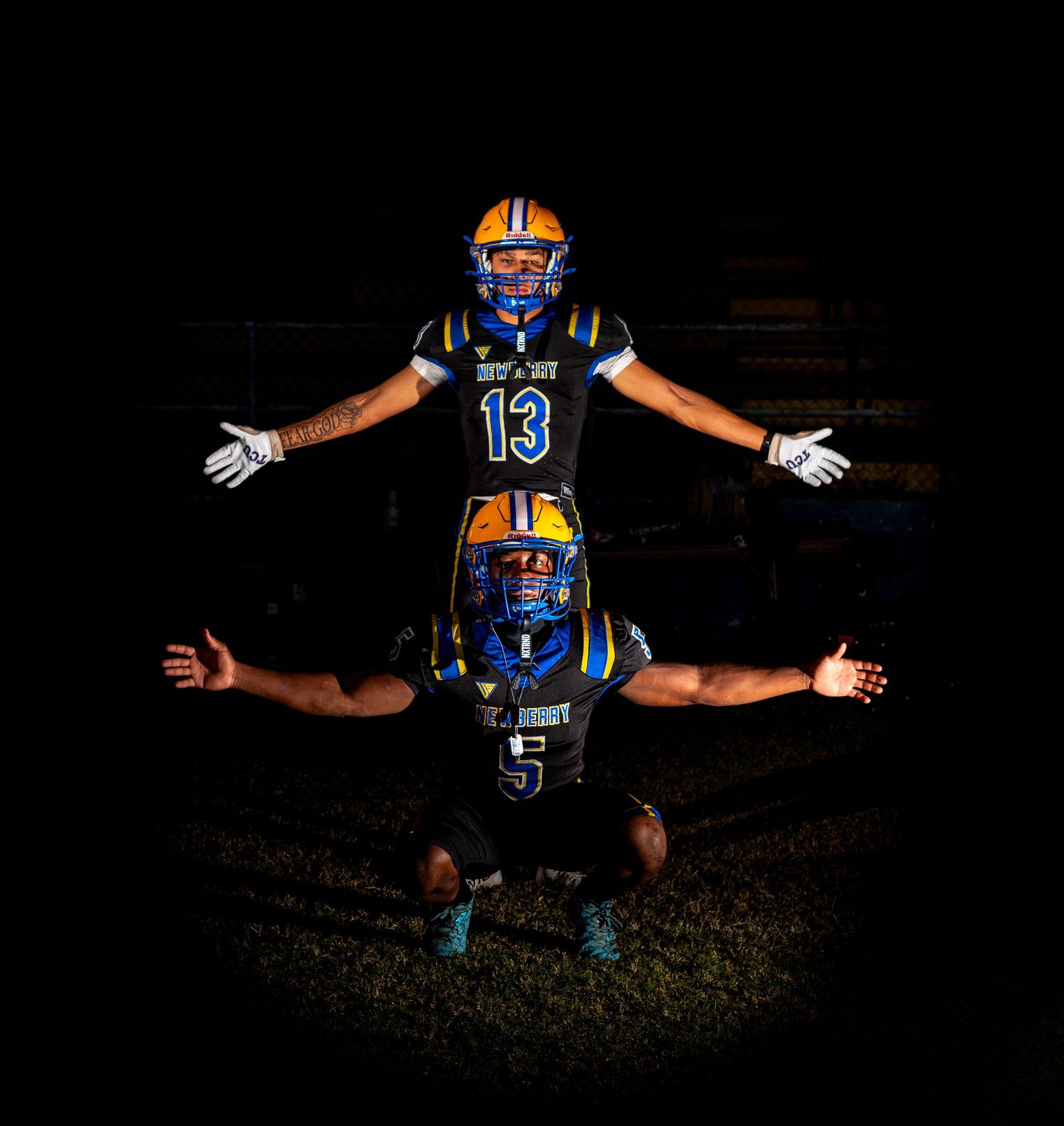 Two football players in blue and gold uniforms pose dramatically under stadium lights, one standing with arms wide, number 13.