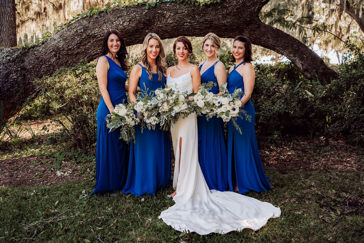 Wedding party in royal blue dresses and white gown poses together outdoors under Spanish moss covered trees.
