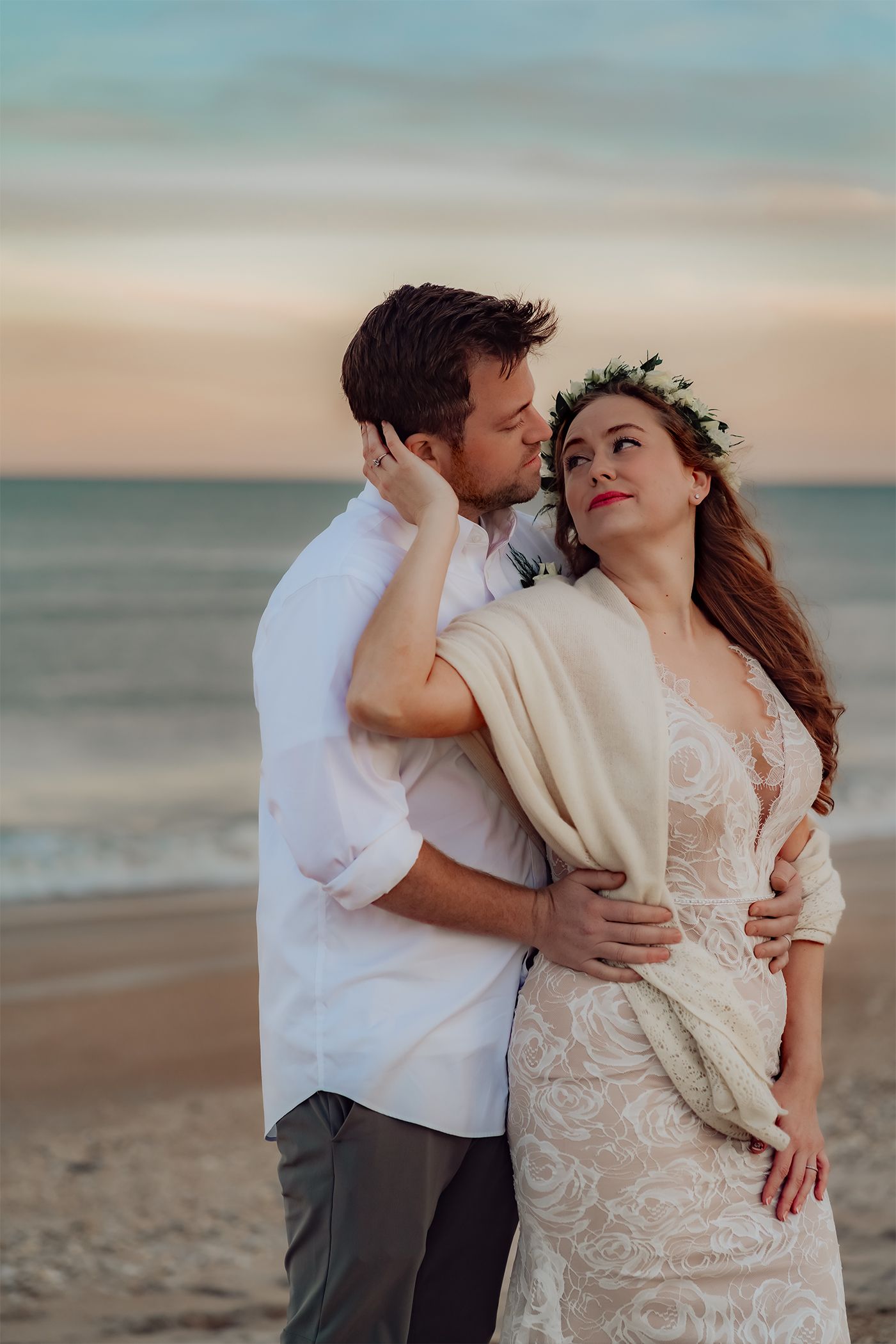 A romantic couple embraces on the beach at sunset, with the bride wearing a lace dress and floral crown.