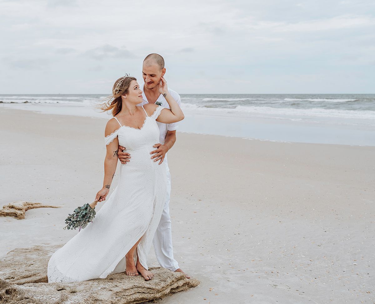 A couple in white attire walks together along a sandy beach with waves and driftwood in the background.