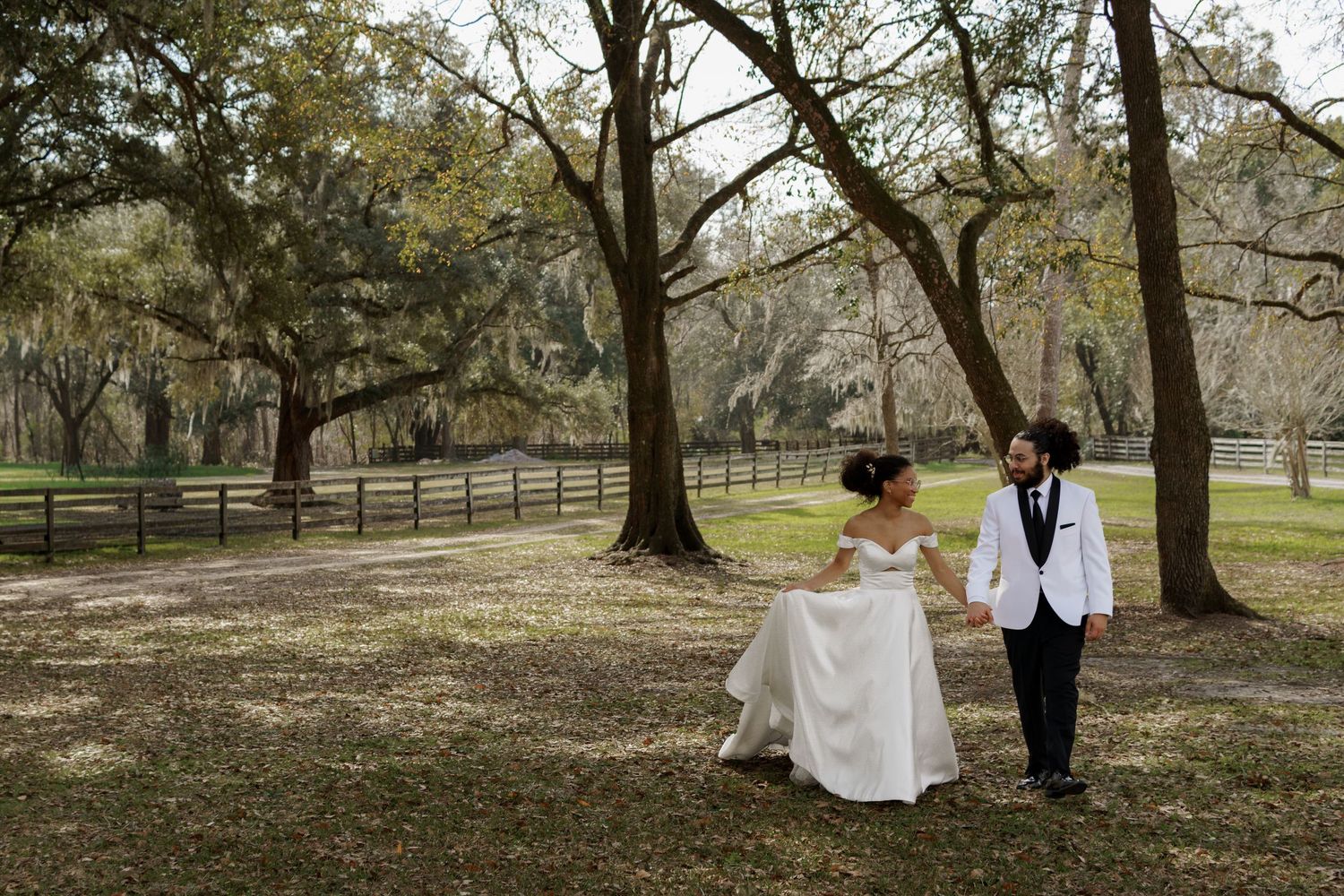 Bride and groom walk hand in hand through a scenic outdoor venue with large oak trees and wooden fence.
