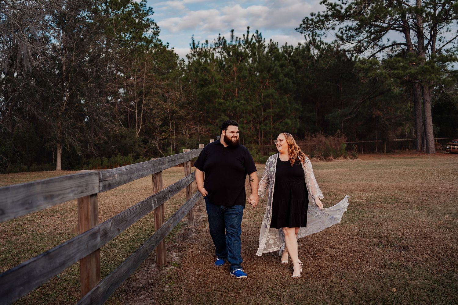 A couple walks along a rustic wooden fence path during a romantic outdoor photo session at sunset.
