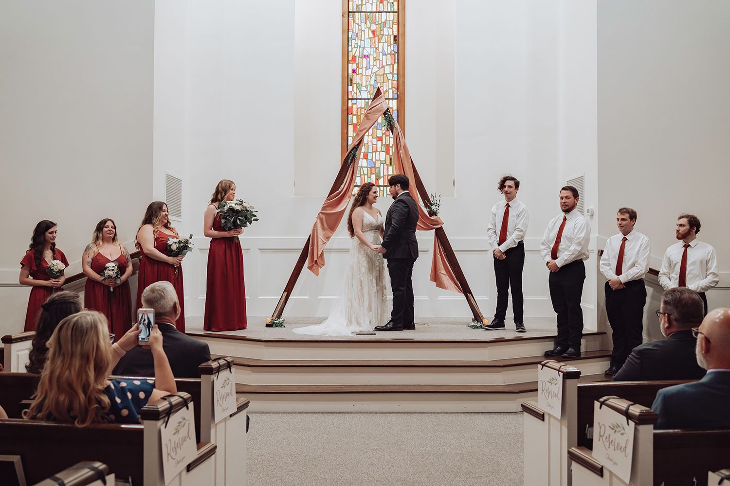 Wedding party members pose in a line with raised arms making victory signs in an elegant white venue.