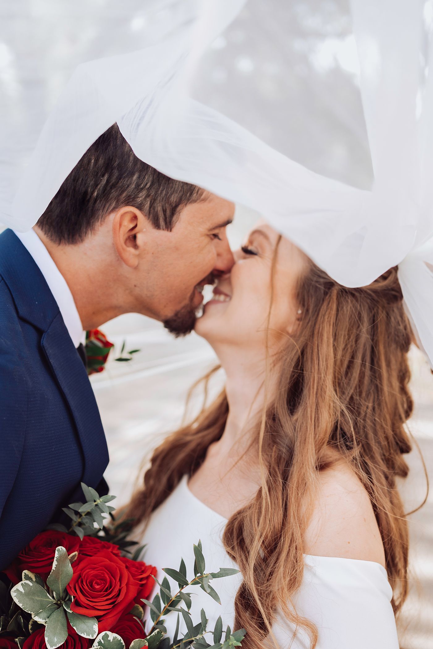 A romantic wedding couple shares tender kisses under a flowing white veil with red roses and greenery.