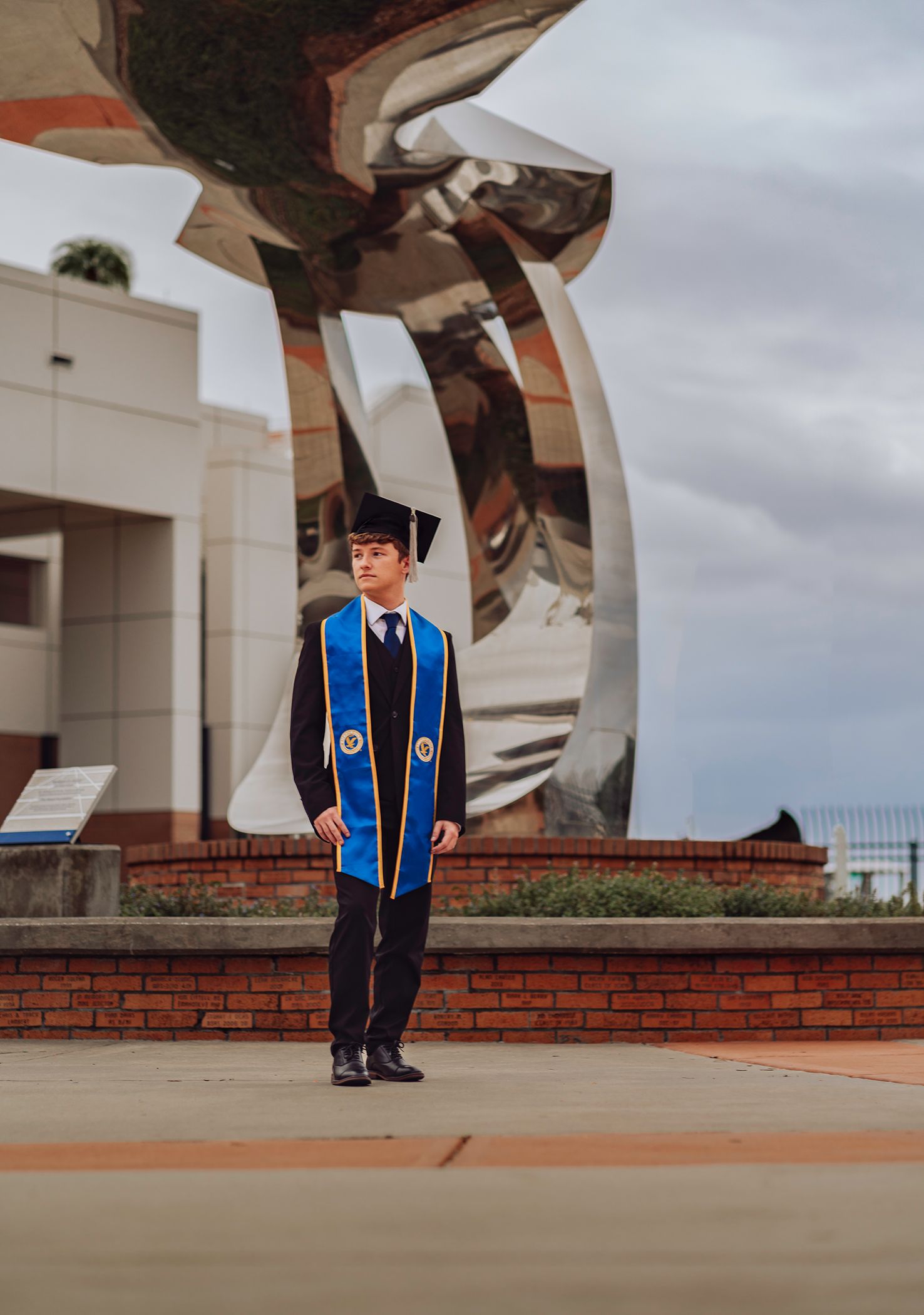 A graduate in blue and black academic regalia stands on a brick terrace during a cloudy graduation day photoshoot in Daytona Beach, FL.