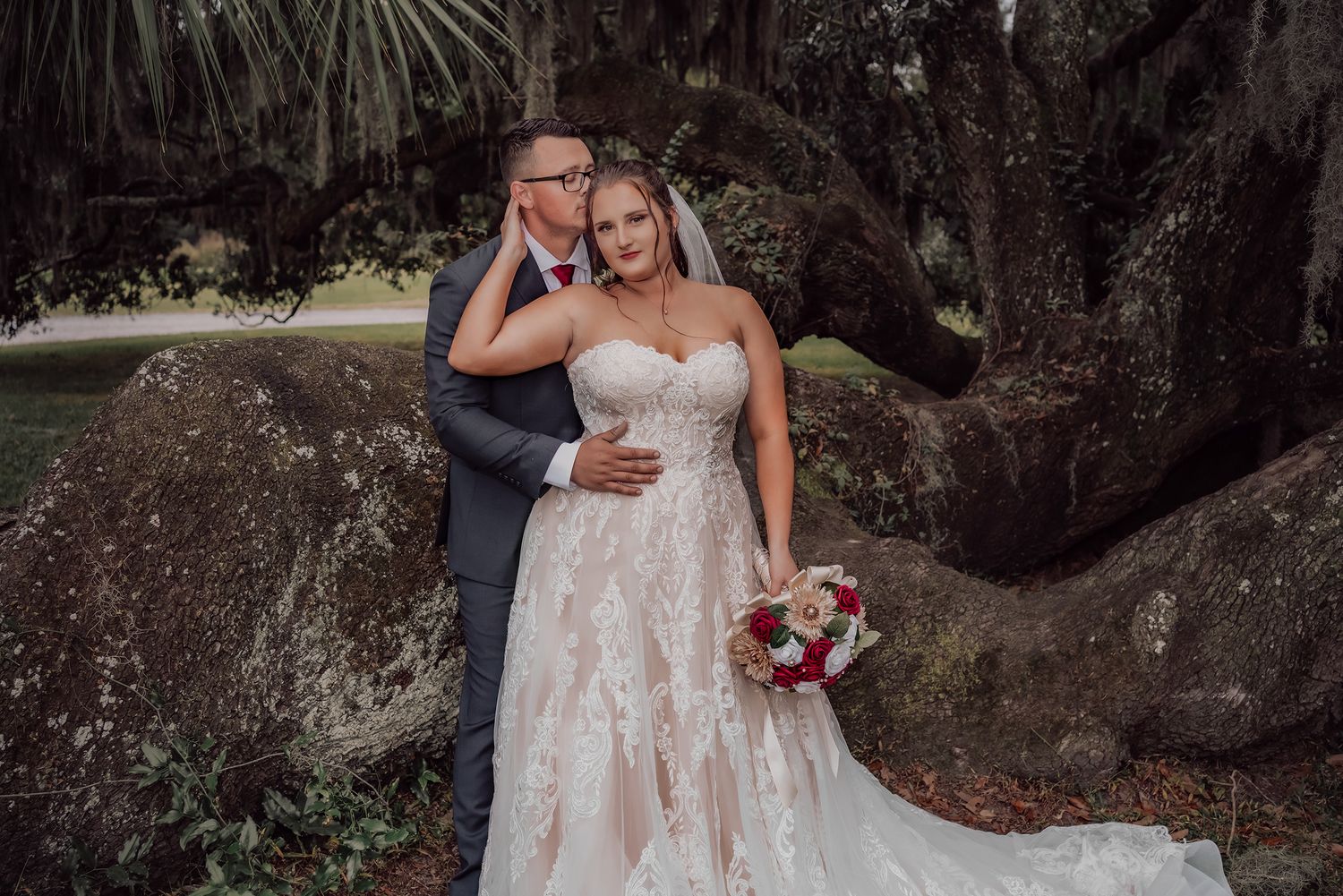 A romantic wedding couple poses beneath towering oak trees draped with Spanish moss at sunset.
