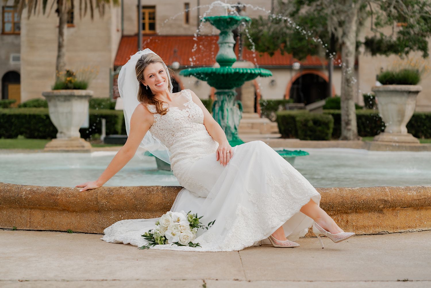 A bride in a white wedding dress sits gracefully by a decorative turquoise fountain in an outdoor setting.