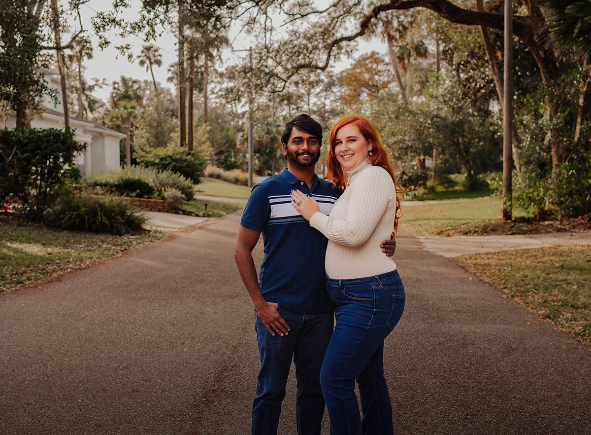 A smiling couple poses on a driveway surrounded by lush trees, the woman with red hair and the man in a blue shirt.