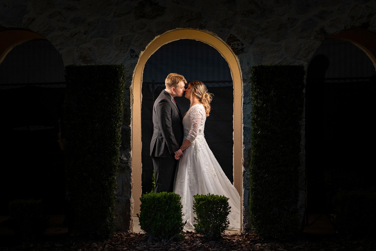 A romantic wedding couple poses in a series of arched doorways at night with dramatic lighting and shadows.