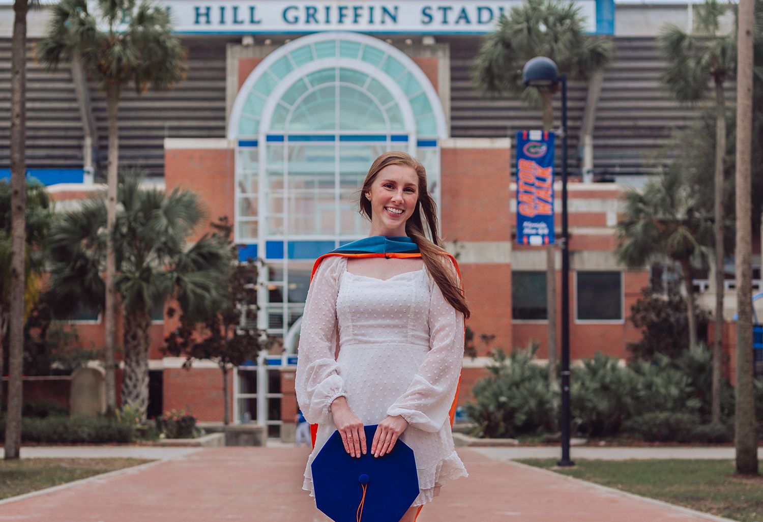 Graduates pose for photos in front of Hill Griffin Stadium wearing blue graduation gowns and casual attire at University of Florida