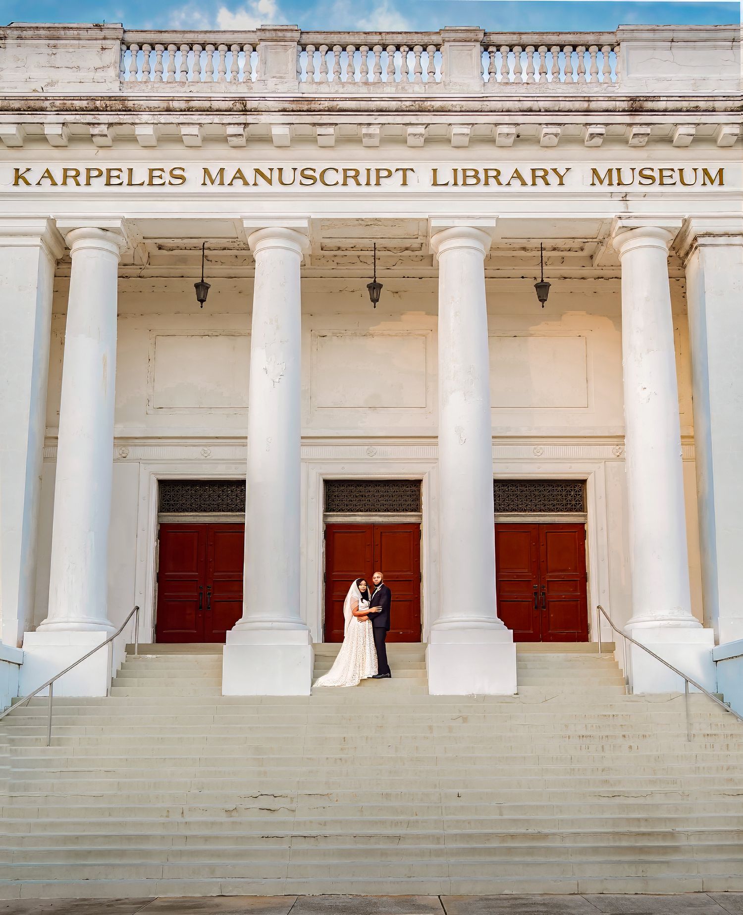 A couple stands between grand white columns at the entrance of the Karpeles Manuscript Library Museum on a sunny day for wedding photography