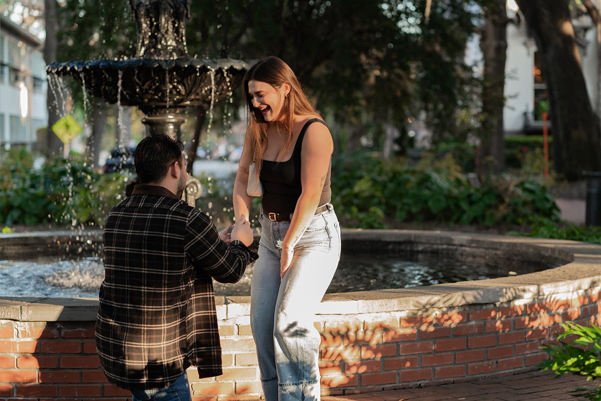 Man proposing to woman near a fountain in a park, she smiles joyfully as he kneels holding her hand.