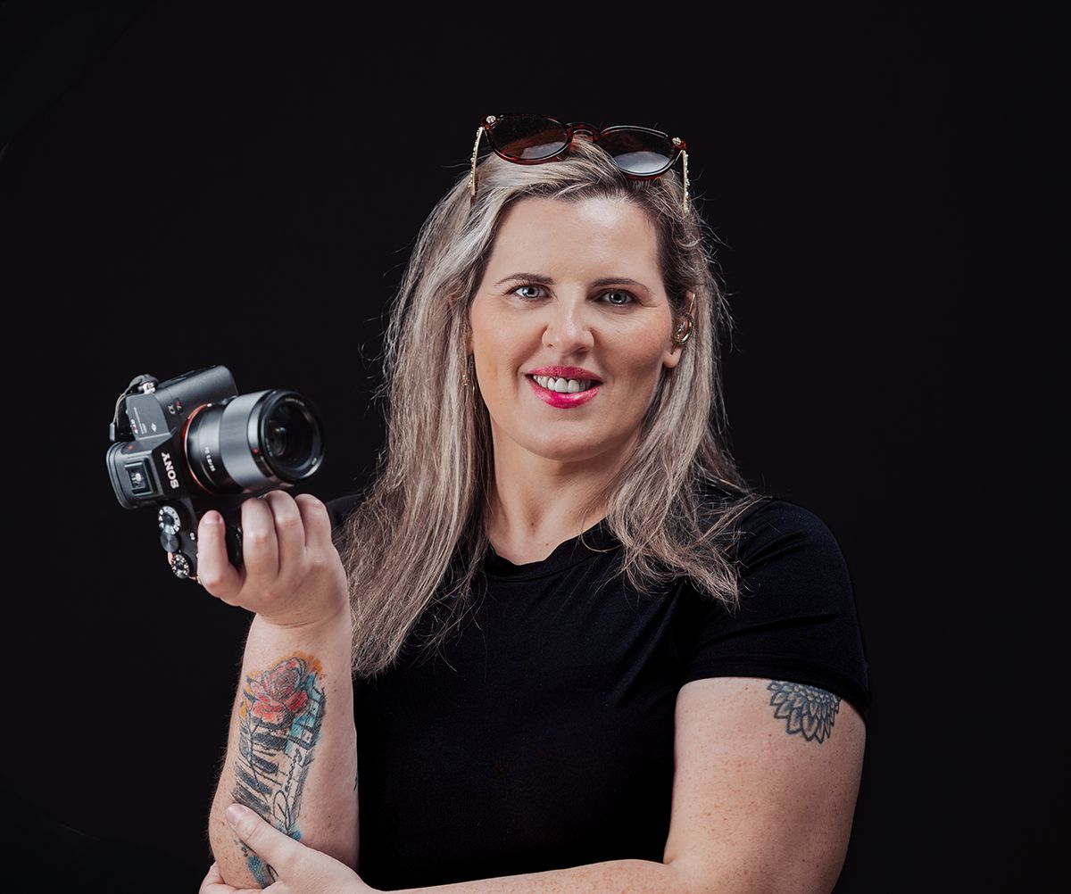 Professional photographer in black shirt holding camera while posing against dark background in studio setting.