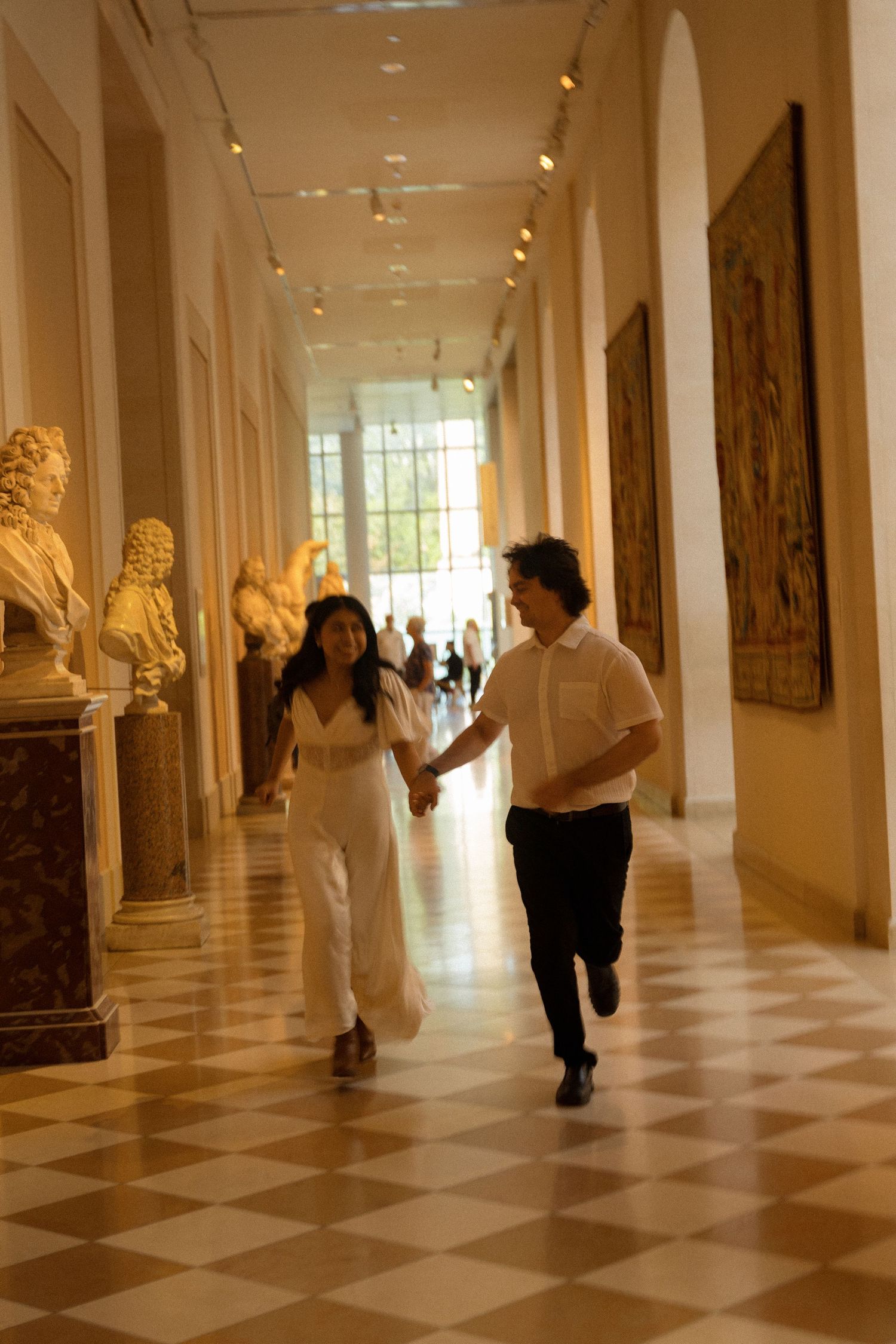 Playful couple running through elegant museum corridor with marble busts and checkered floor.