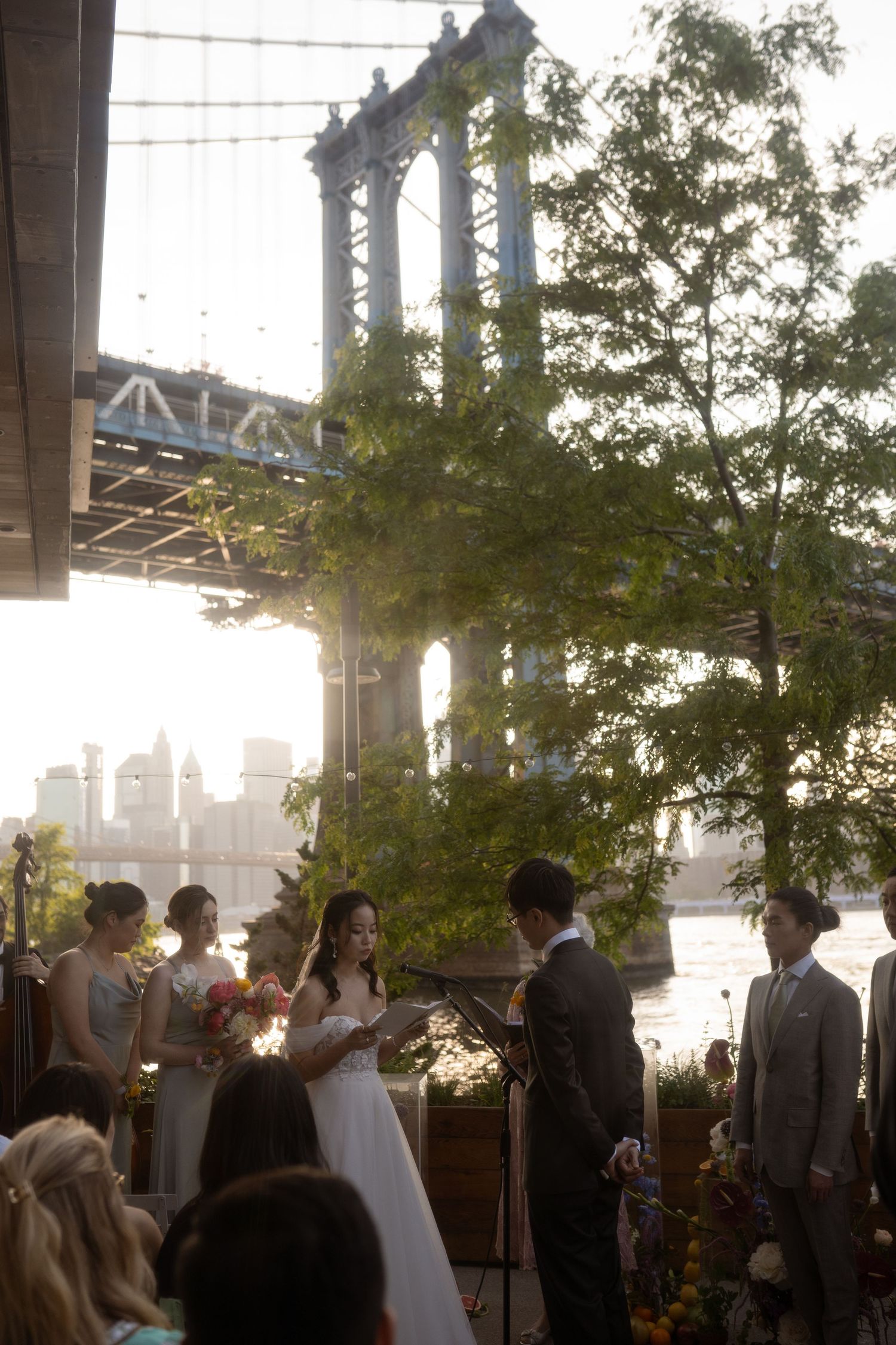 Outdoor wedding ceremony with city skyline visible through string lights at sunset.