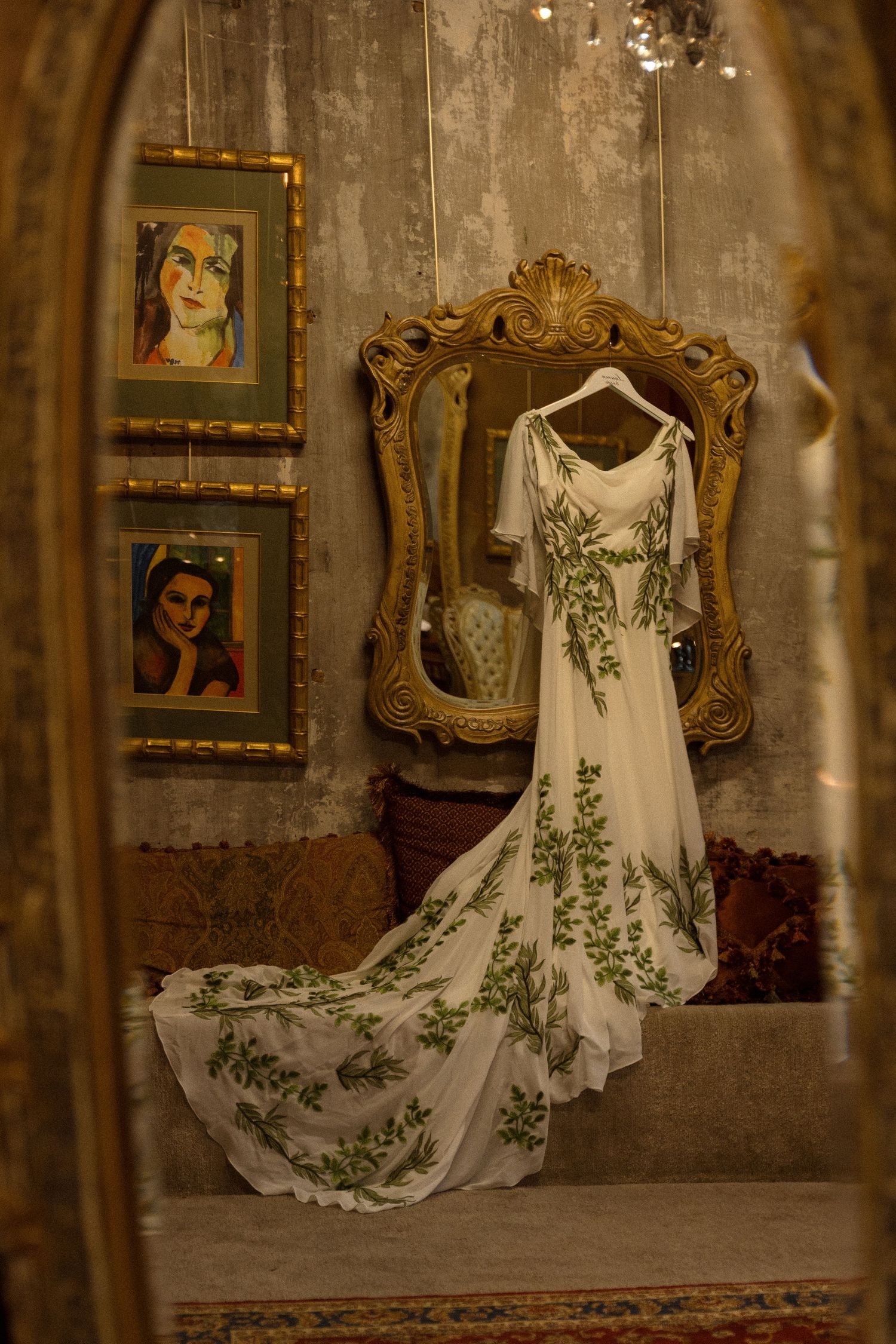 White wedding dress with green floral embroidery displayed against vintage wall with framed portraits.