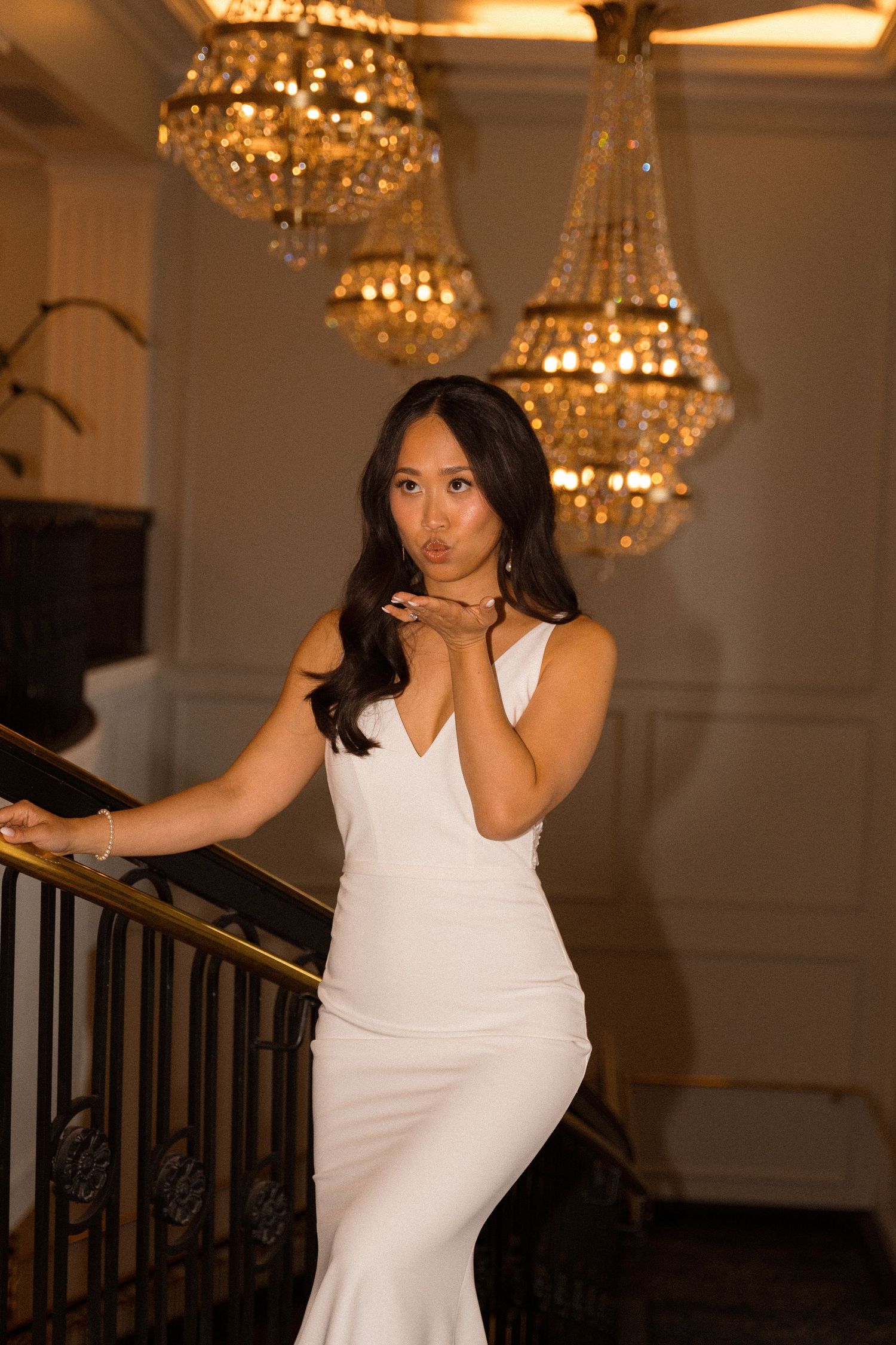 Elegant bride in white dress poses dramatically under ornate chandeliers.