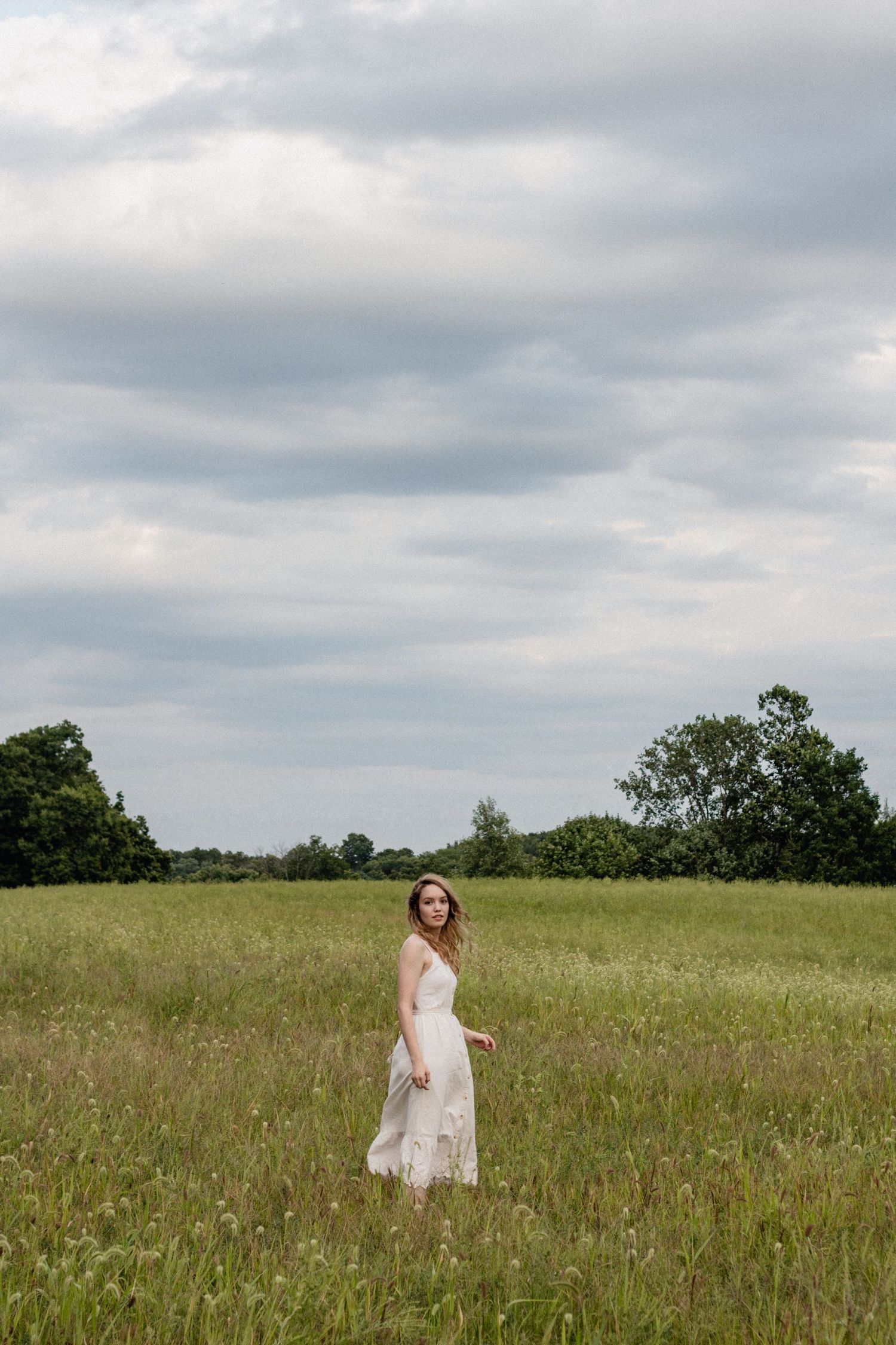 A figure in a white dress walks through a sunlit meadow with tall grass under cloudy skies.