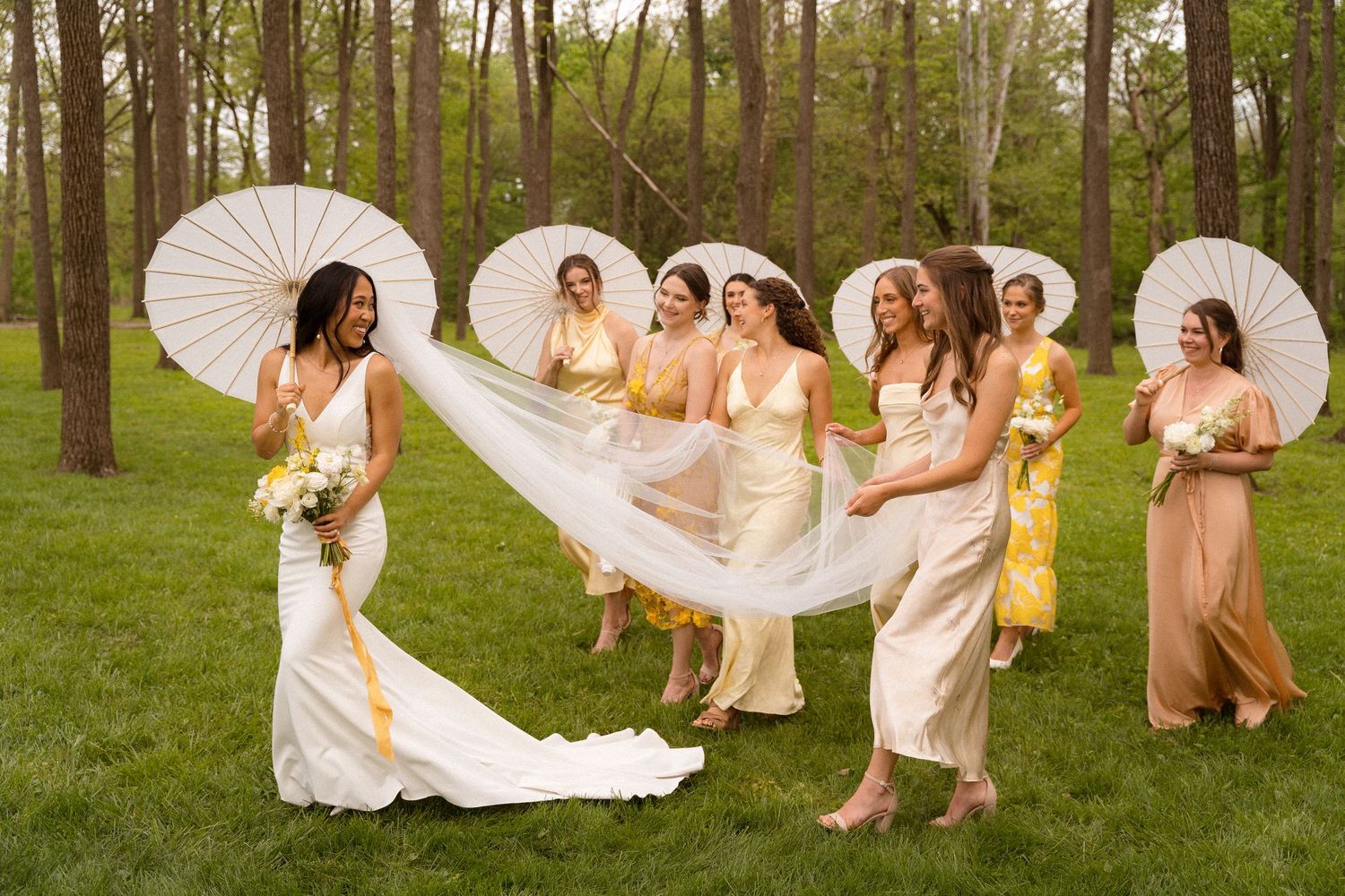 Bridesmaids in champagne dresses help arrange bride's flowing veil outdoors.