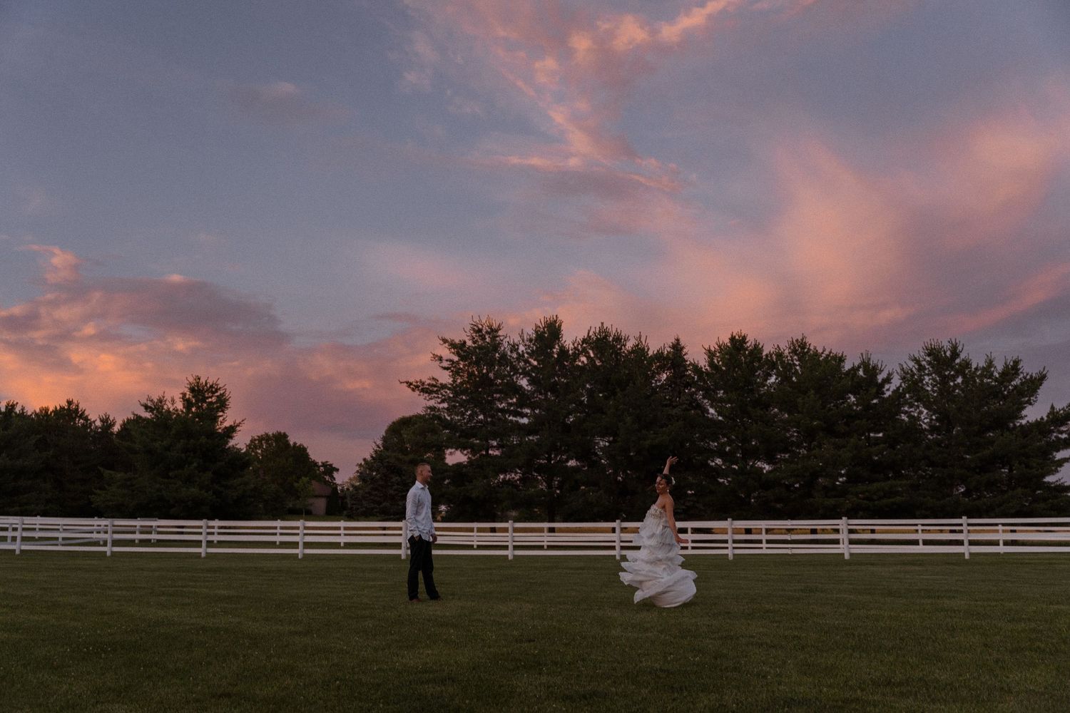 Panoramic sunset view of countryside with white fence and pink sky.