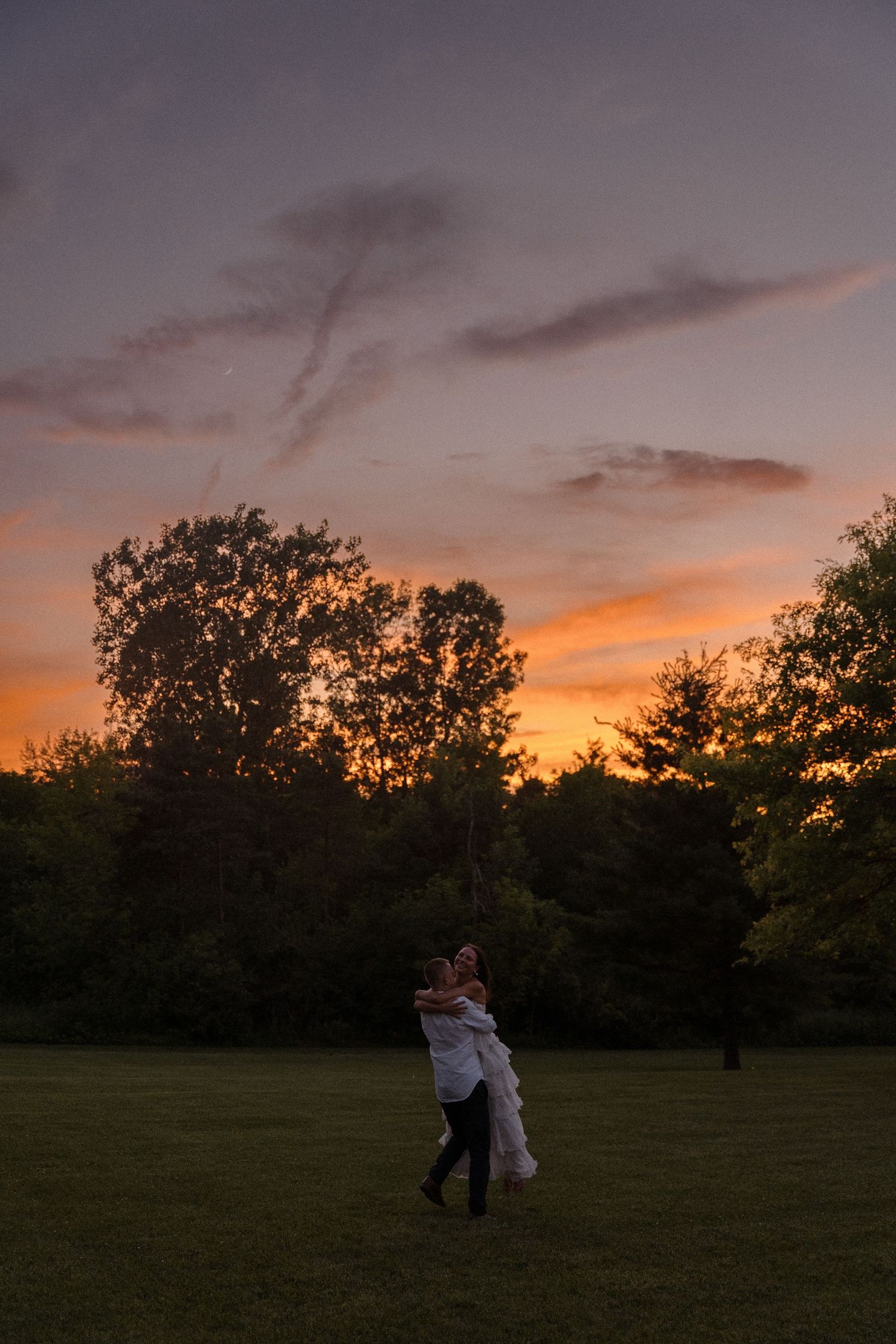 Couple dancing in field silhouetted against dramatic orange sunset.